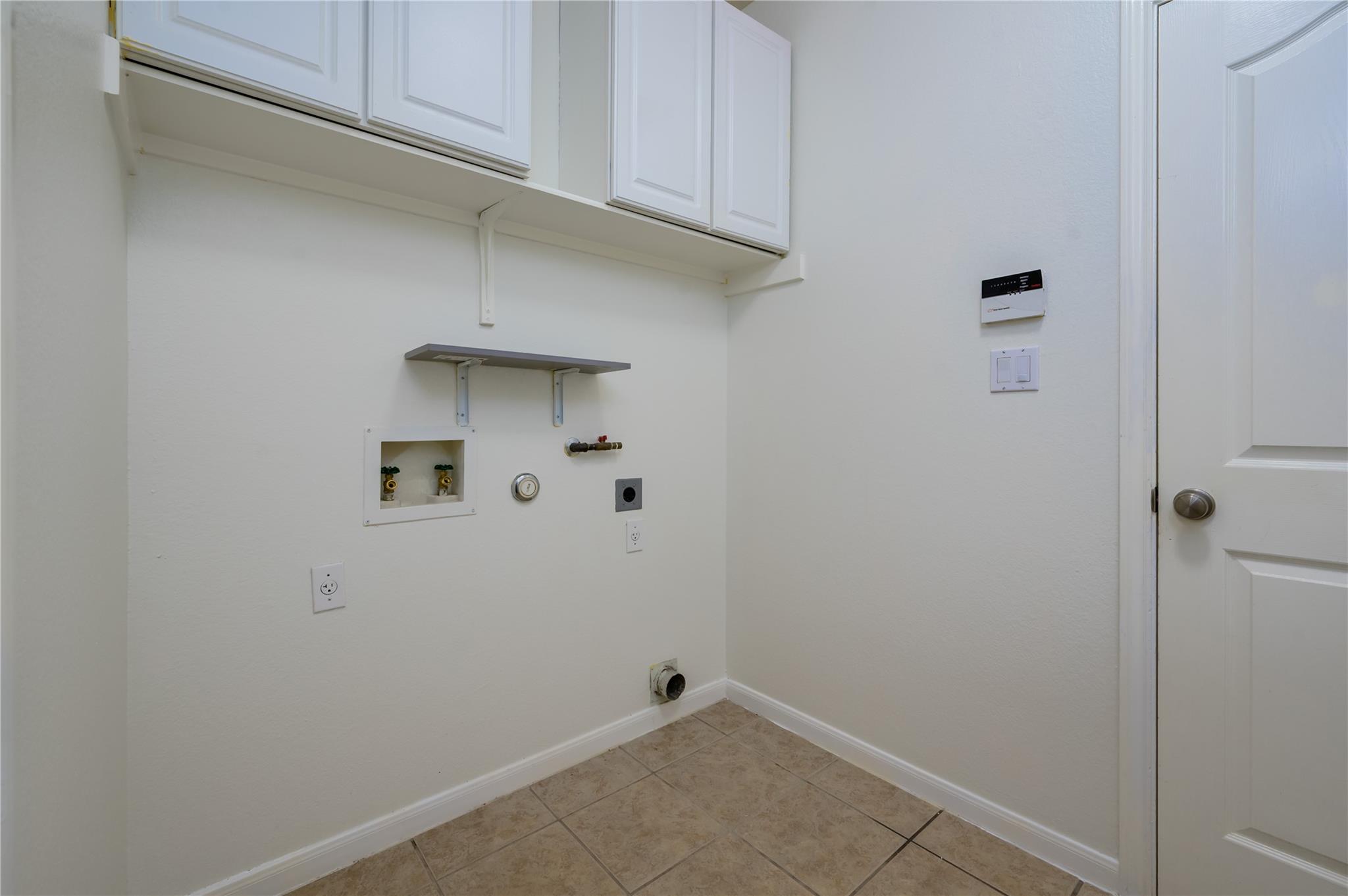 Laundry room with washer hookups, overhead cabinets, and tan tile flooring.