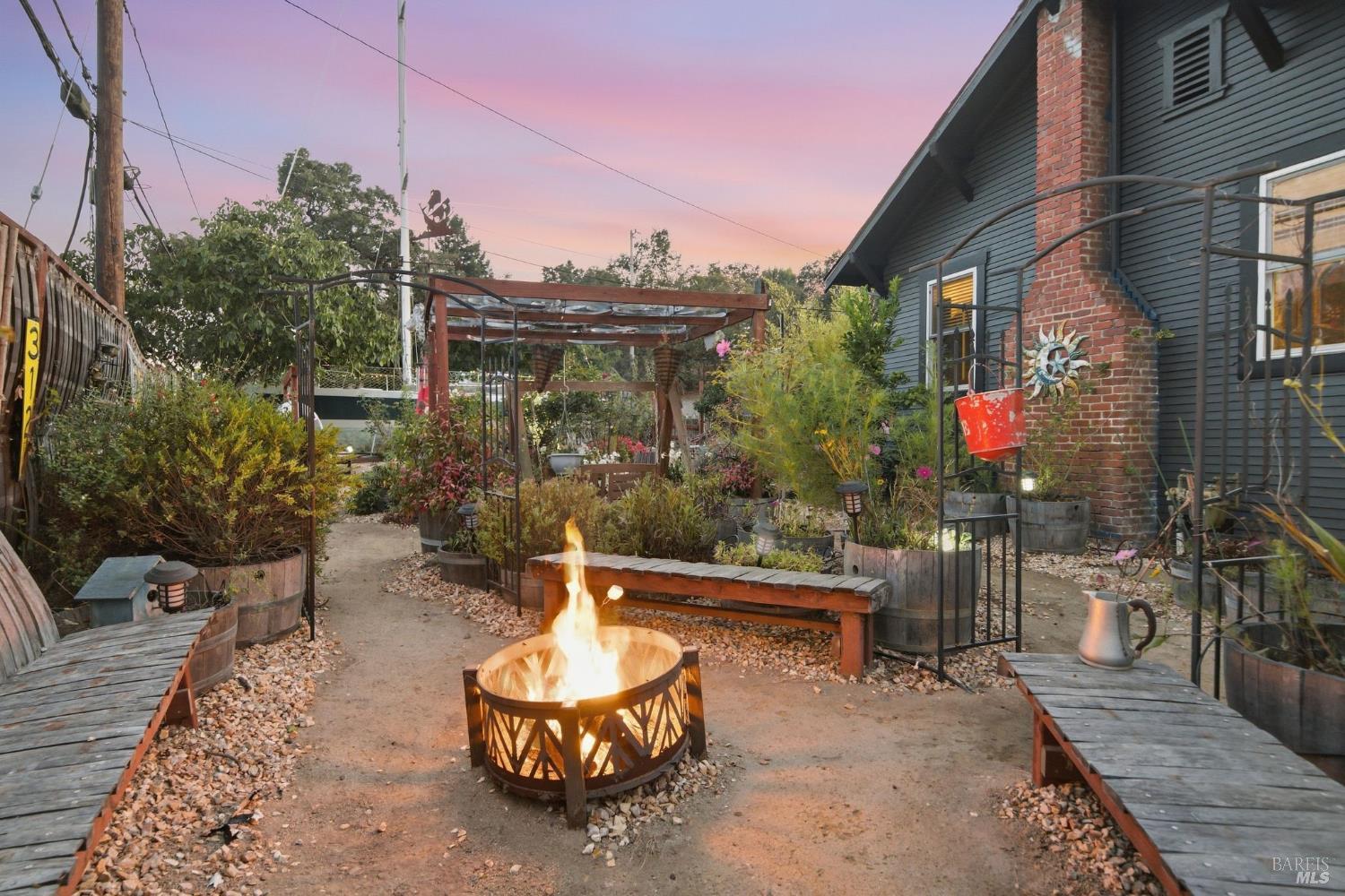 Patio with a glowing fire pit, wooden benches, raised planters, and a brick-chimney house at dusk.