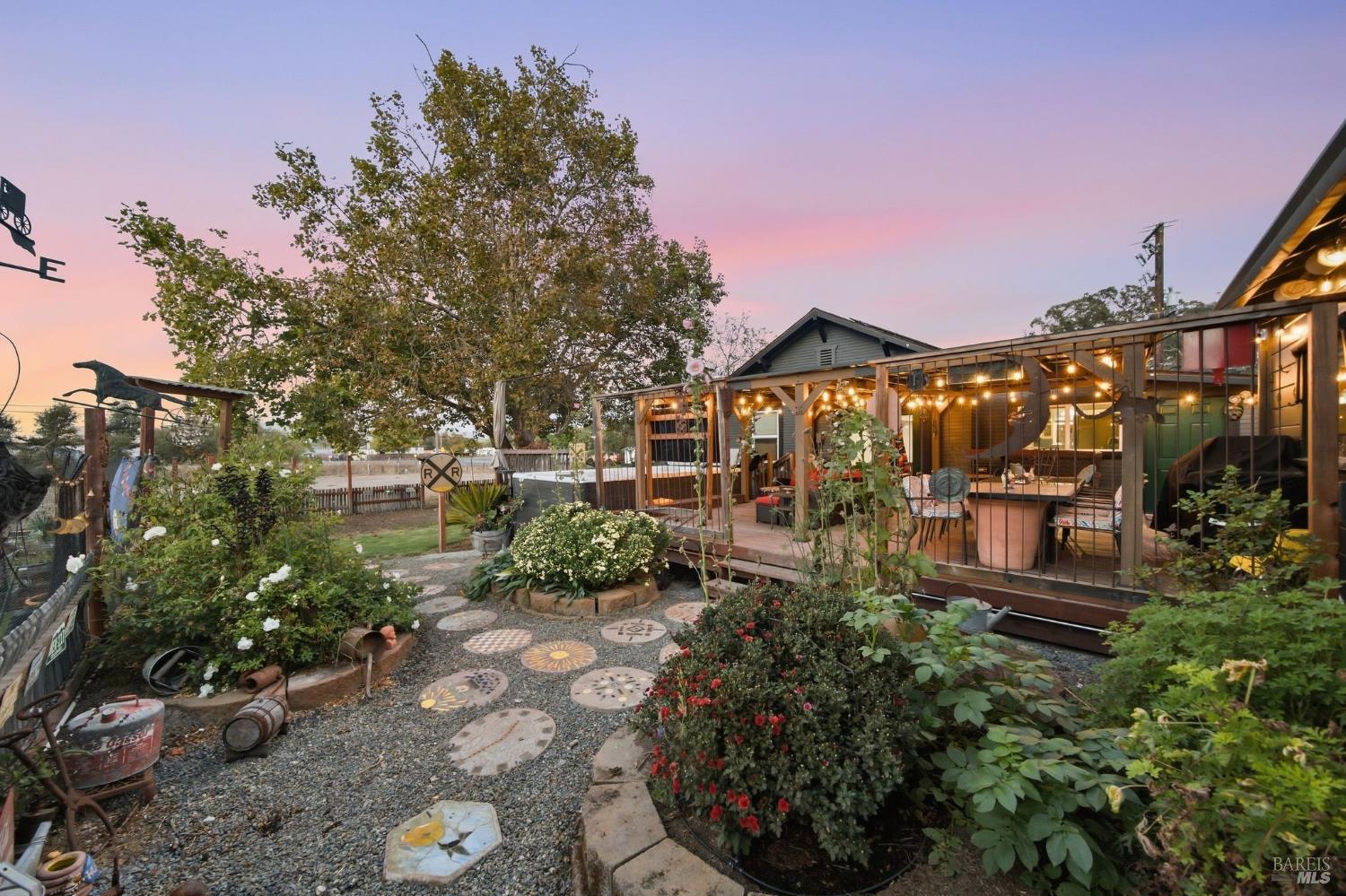Garden patio with string-lit covered deck, gravel paths, flower beds, and a sunset sky.