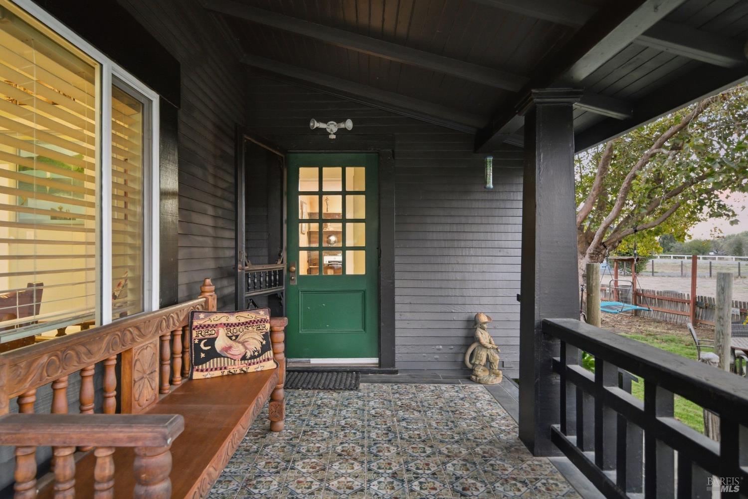 Covered porch with a green entry door, patterned tile floor, and ornate wooden bench overlooking a fenced yard.