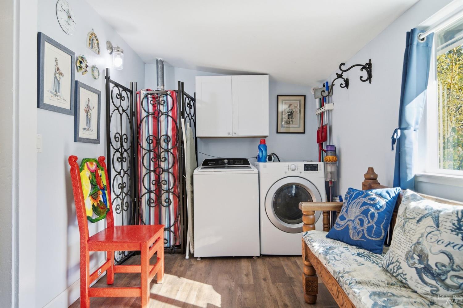 Laundry room with a washer and dryer, white cabinets, and a patterned wooden sofa beside a sunny window.
