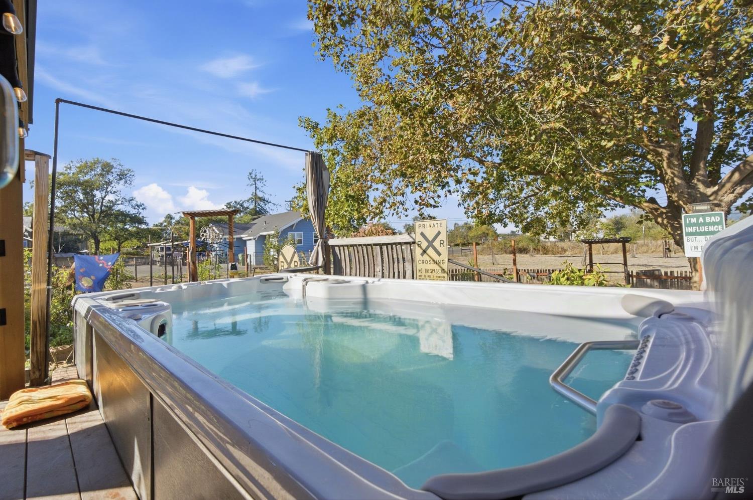 Outdoor hot tub on a wooden deck beneath a large tree, with a fenced yard and blue house beyond.