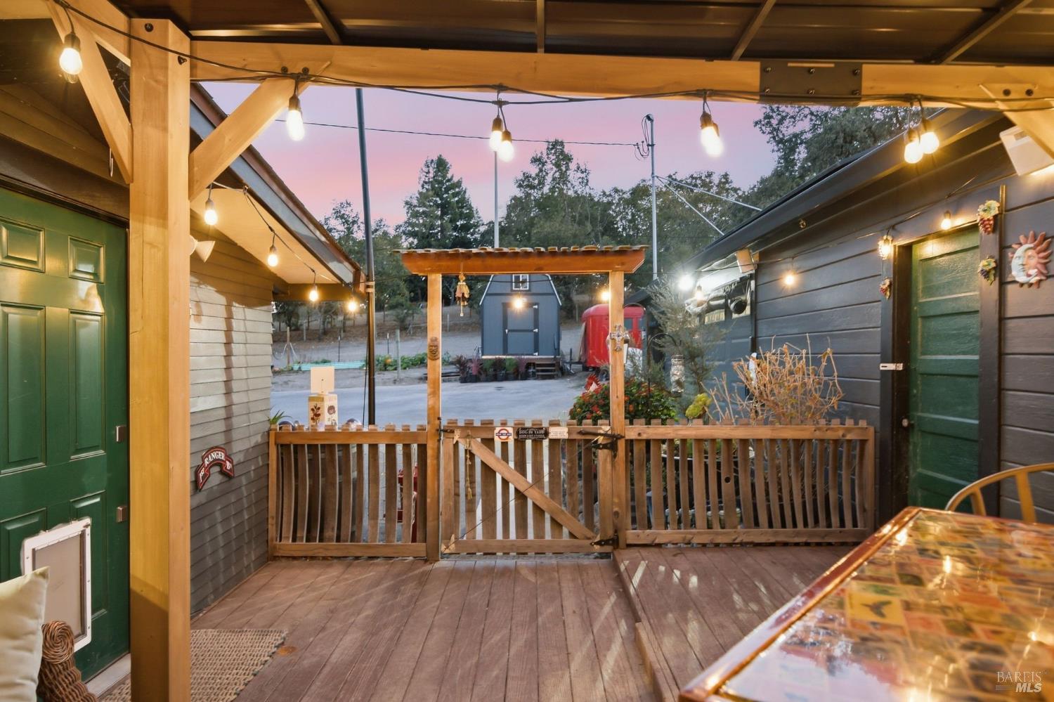 Covered wooden deck with green doors, a fenced seating area, and glowing string lights at dusk.