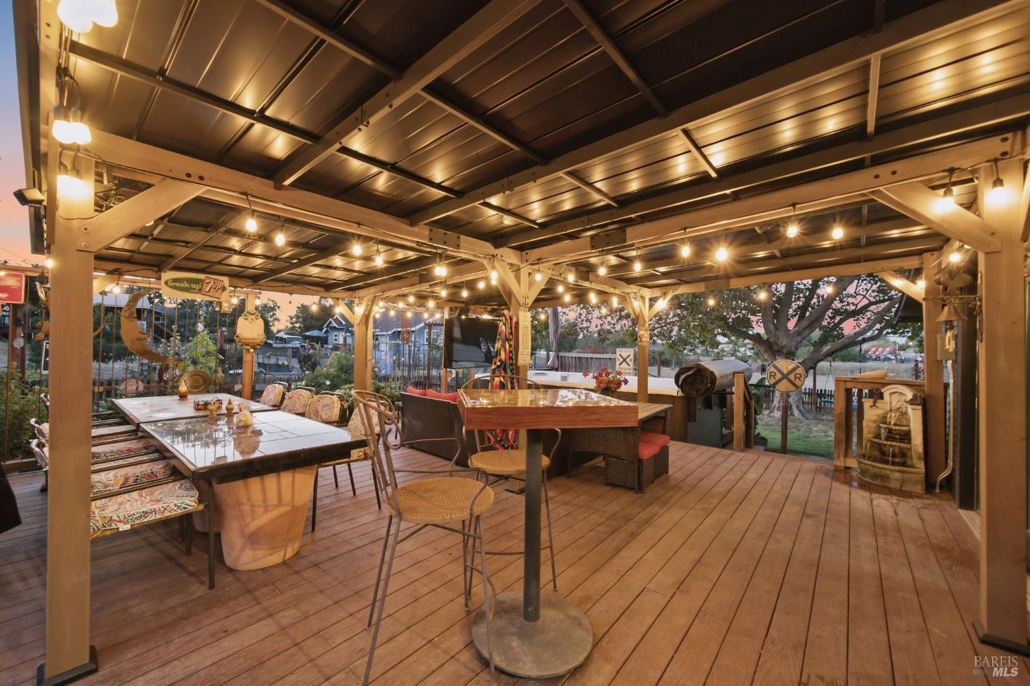 Covered outdoor patio with wooden decking, dining tables, and warm string lights.