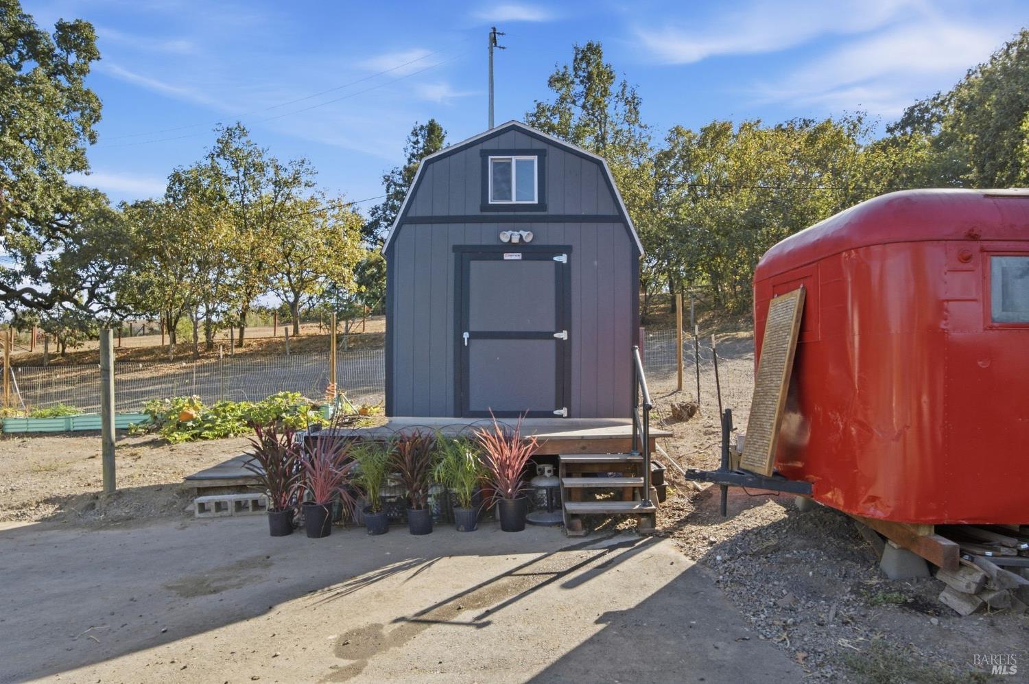 Gray gambrel-roof shed with potted plants at the steps beside a red trailer.