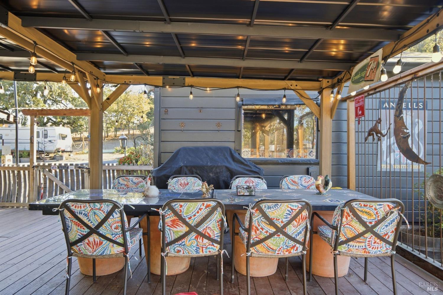 Covered patio with a long dining table, colorful chairs, timber supports, and string lights.