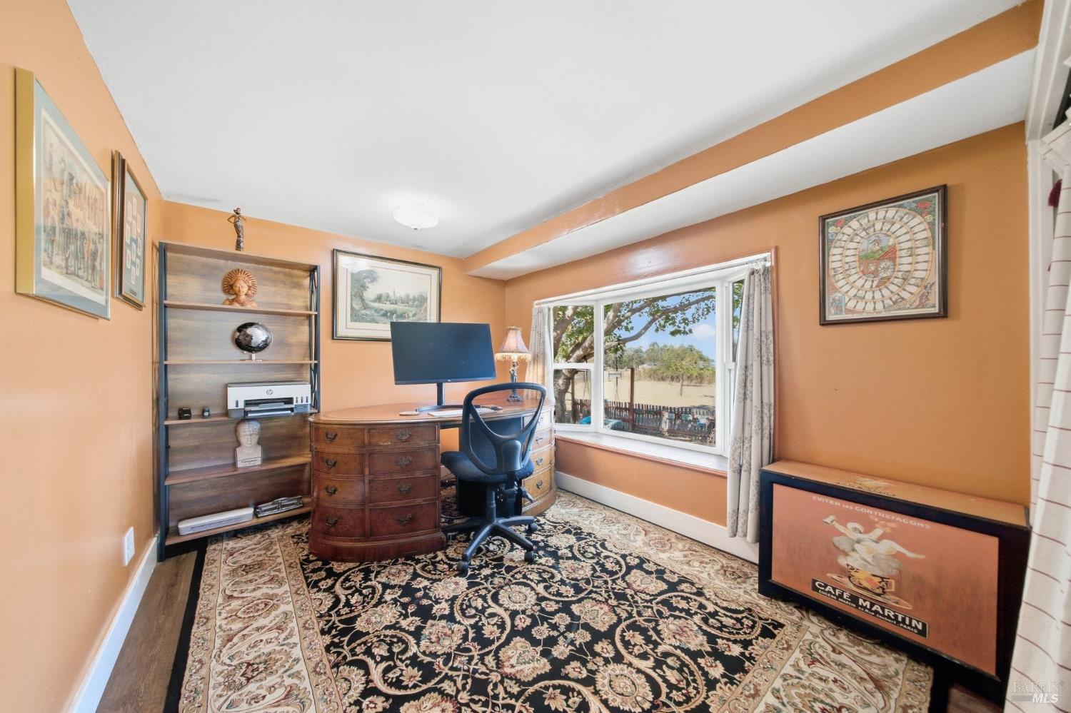 Furnished home office with peach walls, ornate rug, wooden desk, and a window overlooking open land.