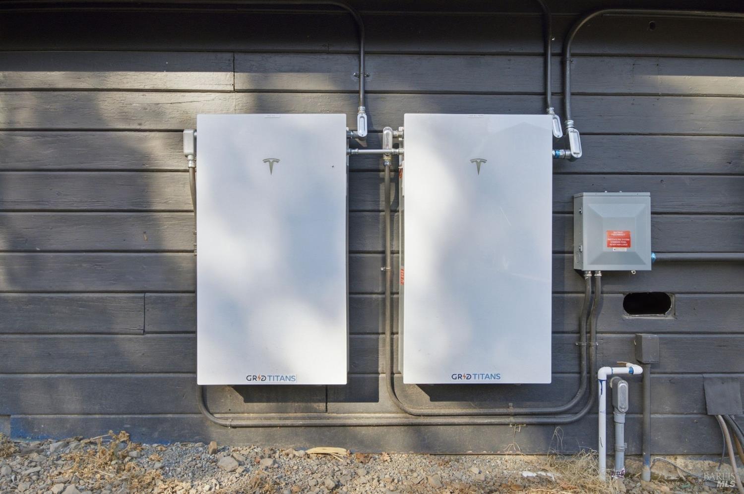 Two Tesla energy storage units mounted on dark exterior siding with exposed conduit.