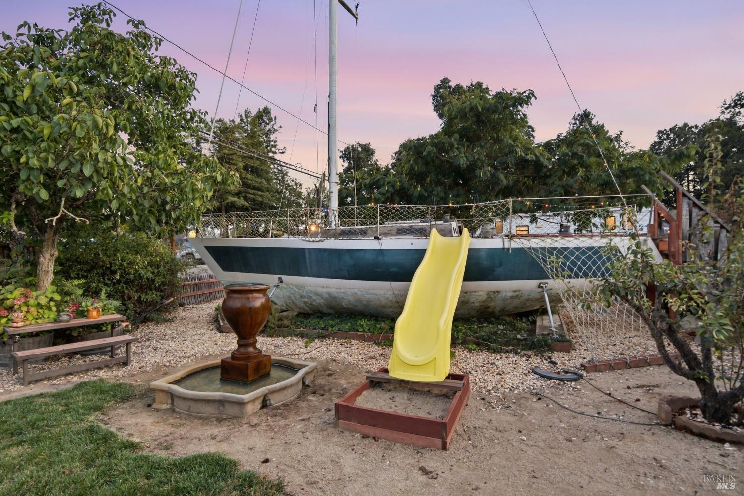Yard with a boat on land, yellow slide, and small fountain beneath a pink dusk sky.