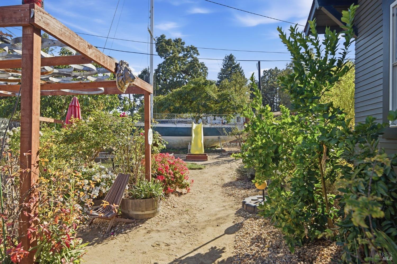 Landscaped yard with a wooden pergola, garden seating, flowering plants, and a boat in the background.
