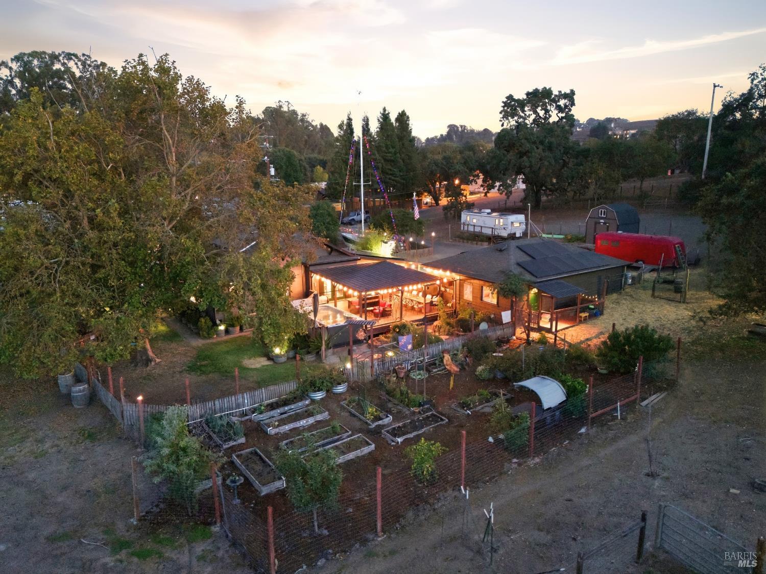 Aerial view of a fenced home and garden at dusk, with a lit deck and solar panels.