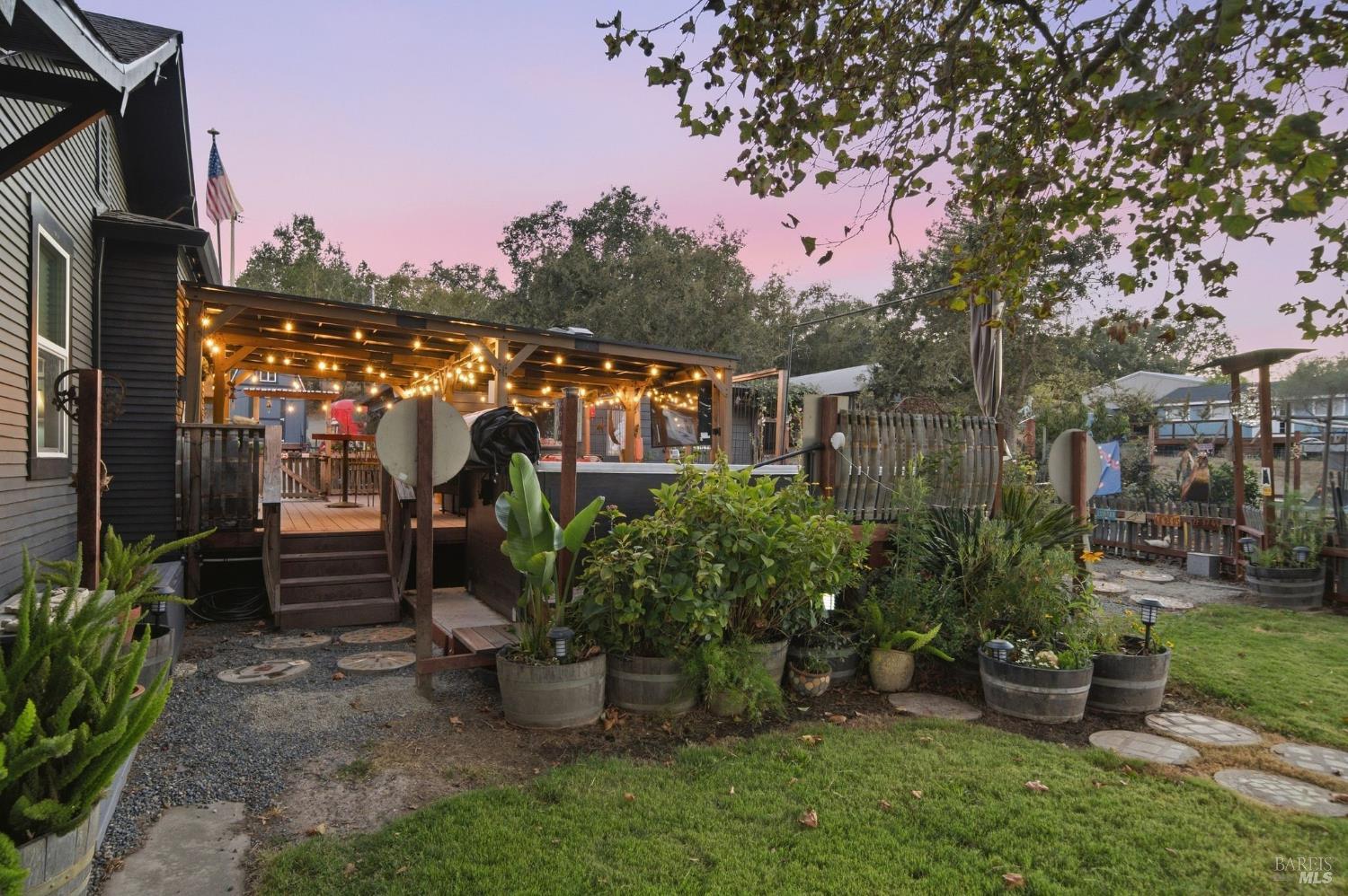Covered backyard deck with string lights, garden beds, and lawn at dusk.