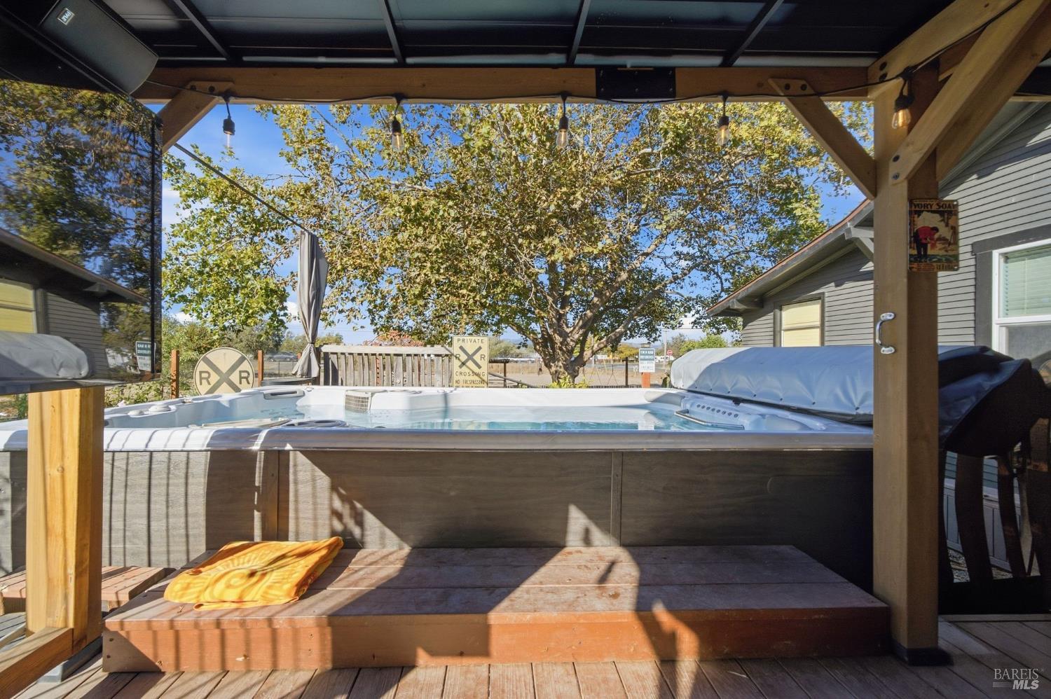 Outdoor hot tub beneath a timber pergola, with a wood deck and leafy tree beyond.