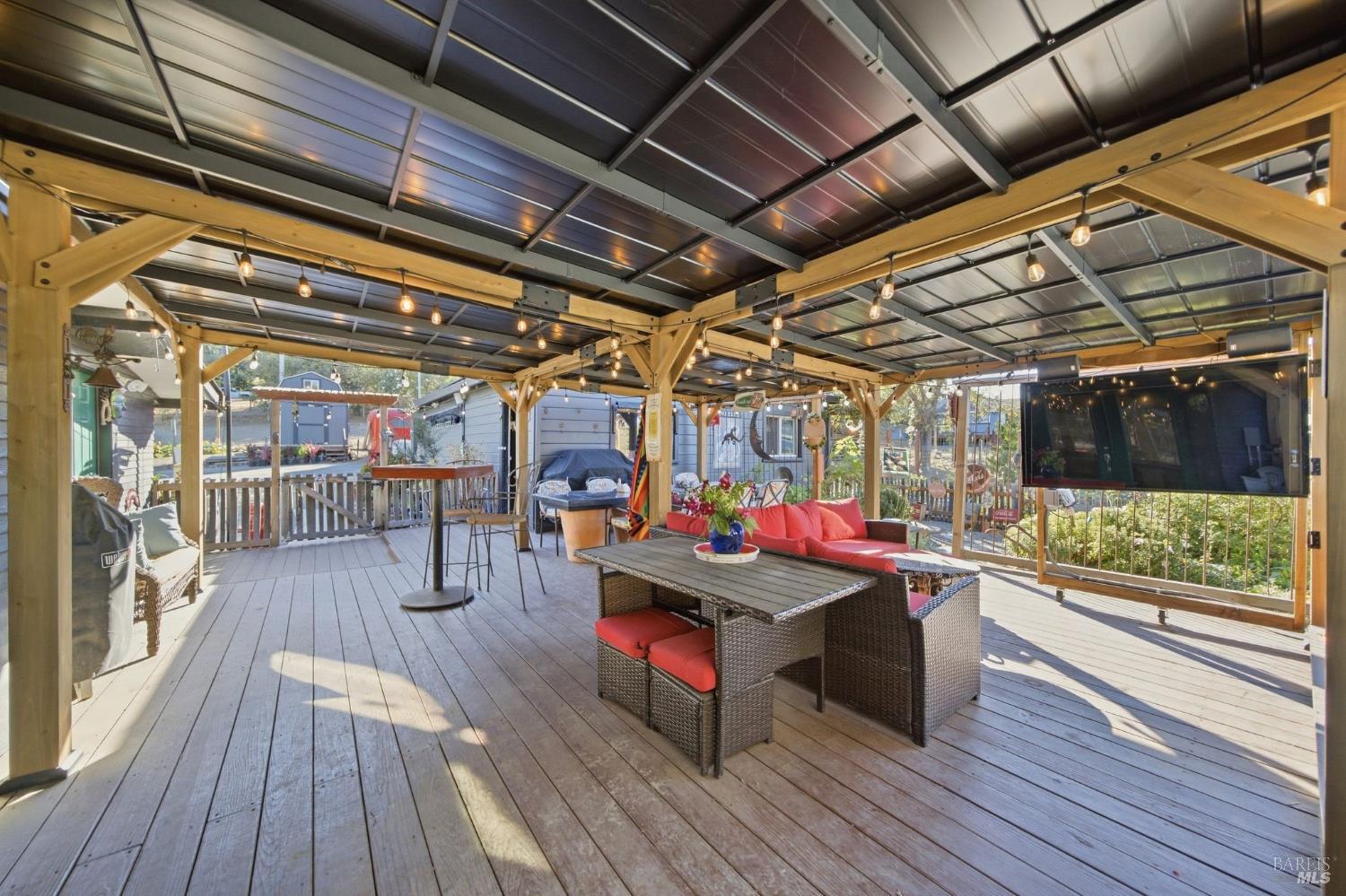 Covered deck with exposed timber beams, string lights, red outdoor sofa, and wicker dining set.
