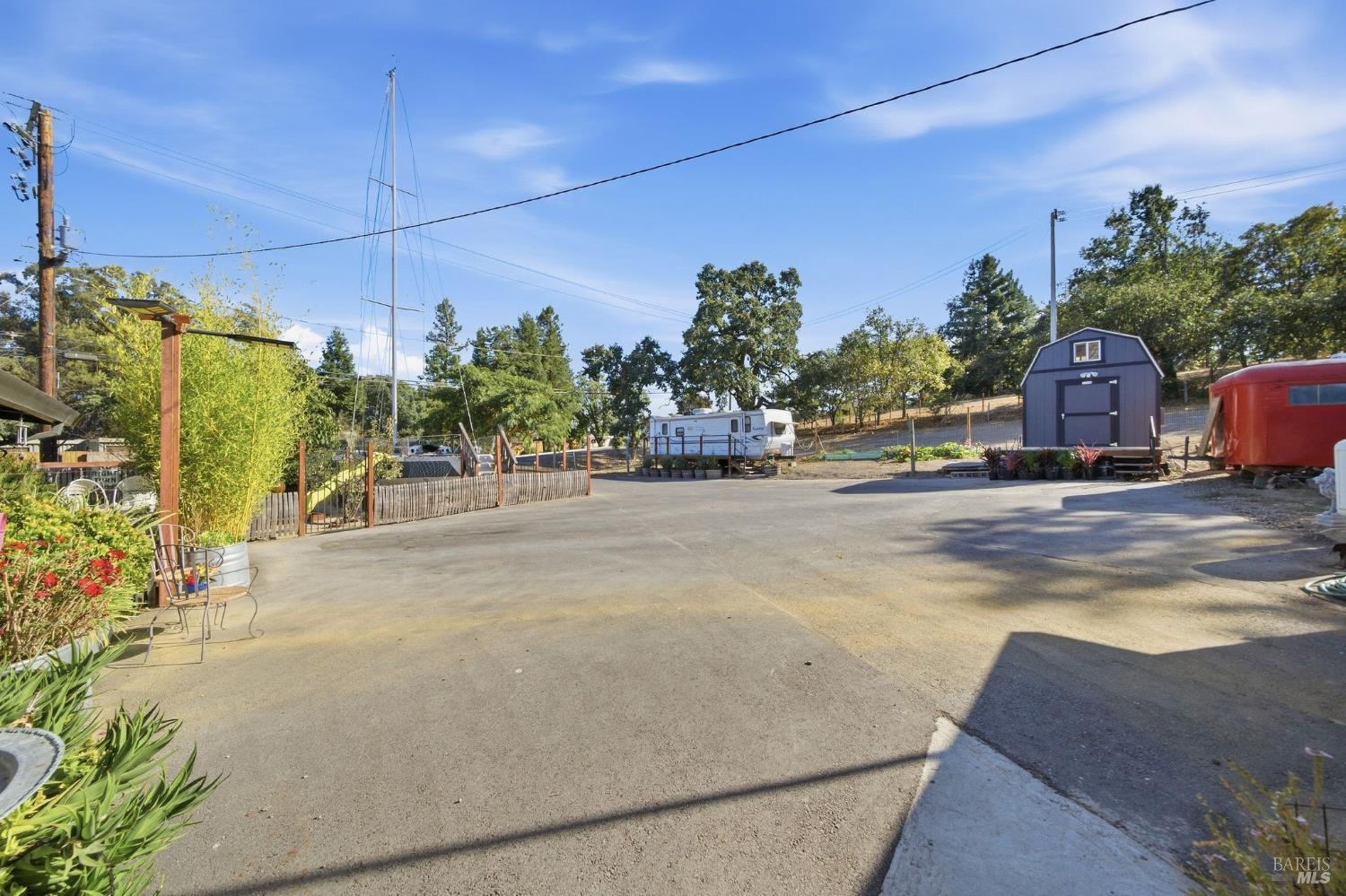 Paved property yard with a dark shed, travel trailer, red trailer, and garden beds.