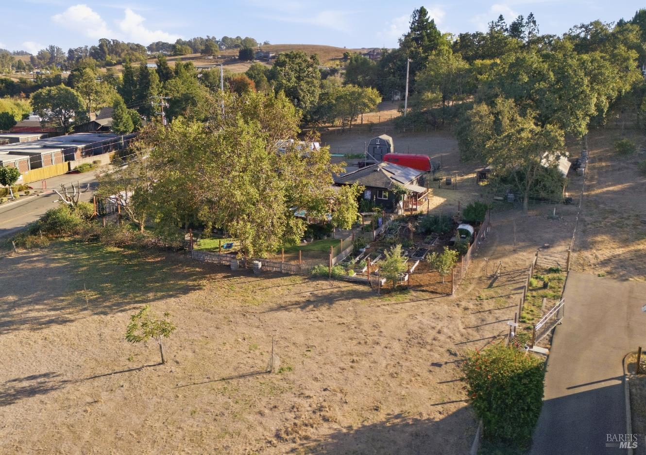 Fenced rural property with a small house, garden beds, and open land viewed from above.