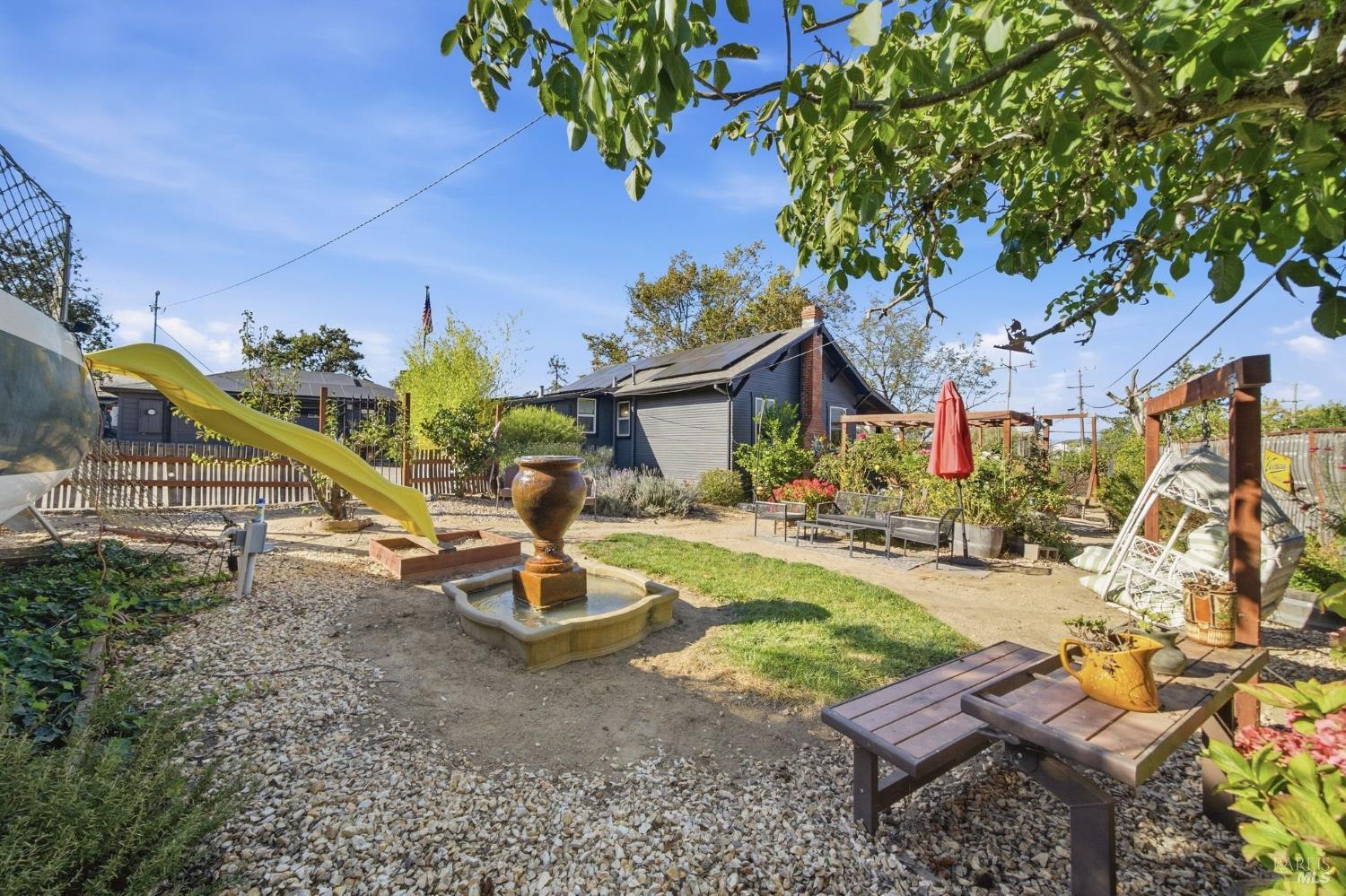 Backyard with a stone fountain, yellow slide, outdoor seating, and a pergola beside a solar-paneled house.