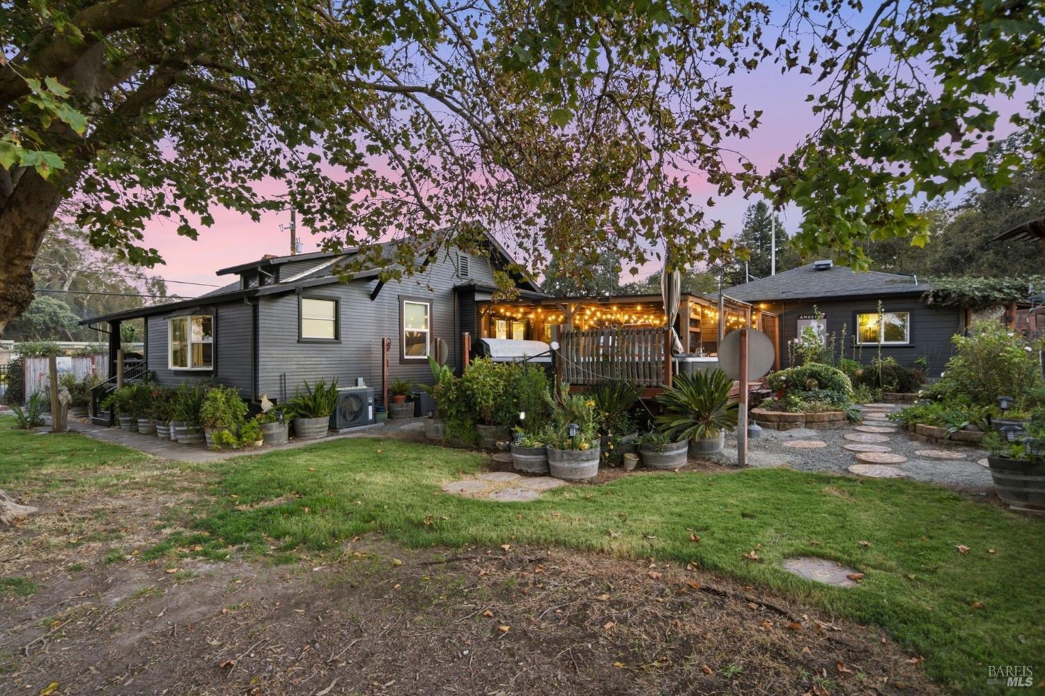 Gray house with a warmly lit covered patio, garden beds, and a stepping-stone path at dusk.