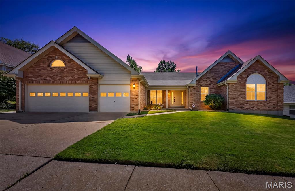 Brick-and-siding home at dusk with two garages, glowing windows, and a broad green lawn.
