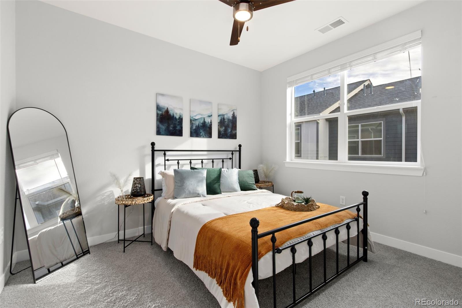 Staged bedroom with a black metal bed, rust-colored throw, large window, and full-length mirror.