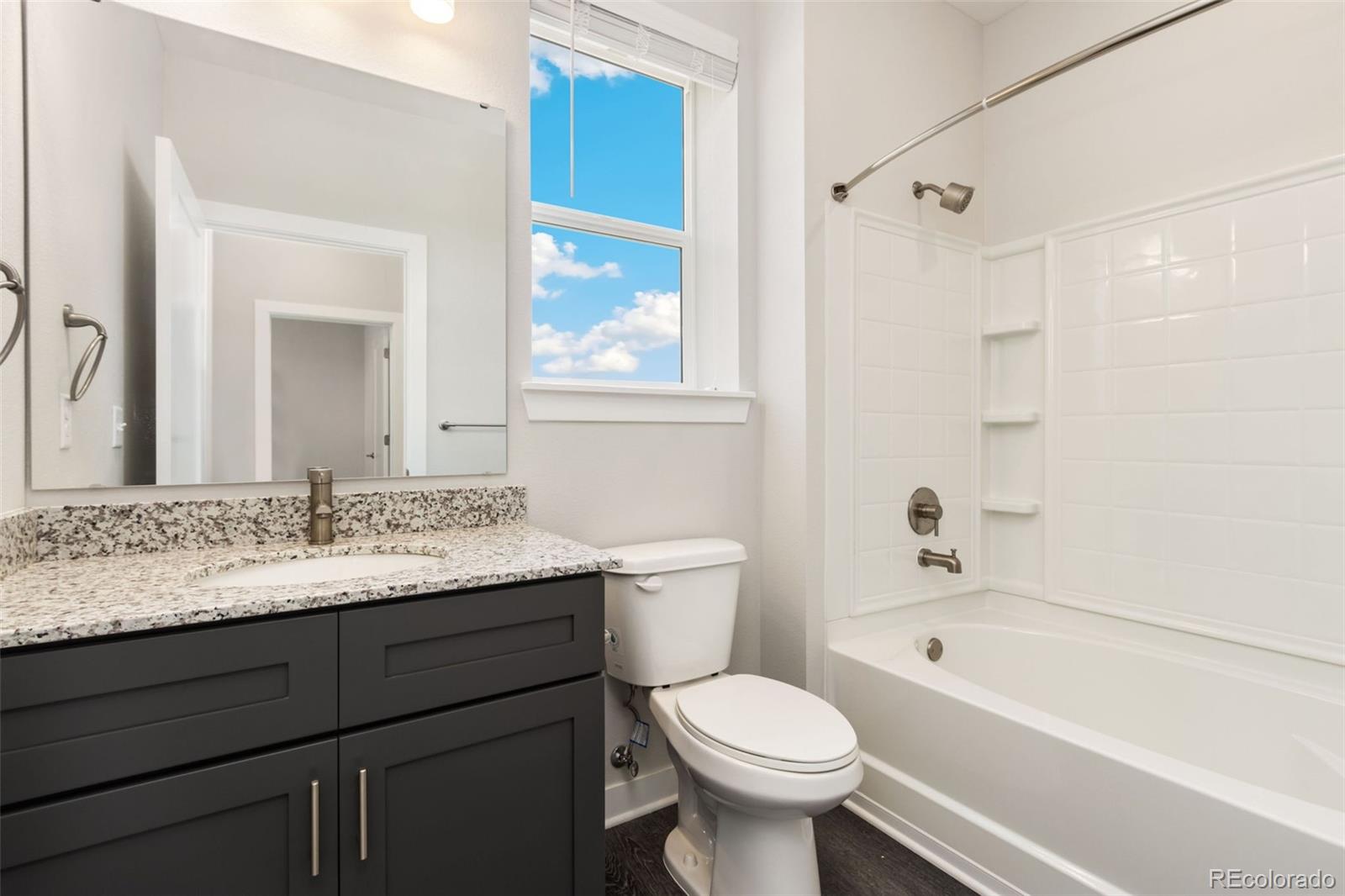 Bathroom with a granite-topped vanity, toilet, and white tub-shower beside a tall window.
