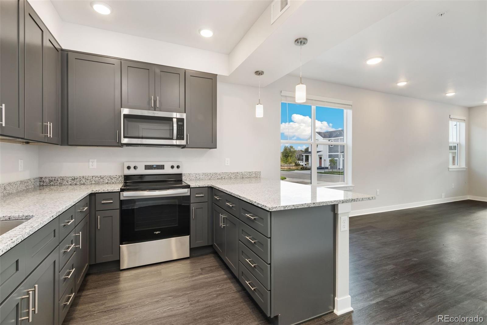 Vacant kitchen with dark gray cabinets, speckled counters, stainless steel appliances, and an adjoining living area.