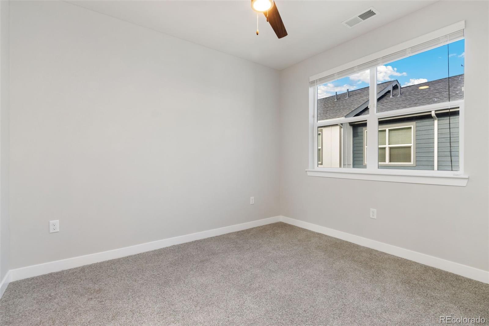Vacant carpeted bedroom with a ceiling fan and a large window overlooking a neighboring building.