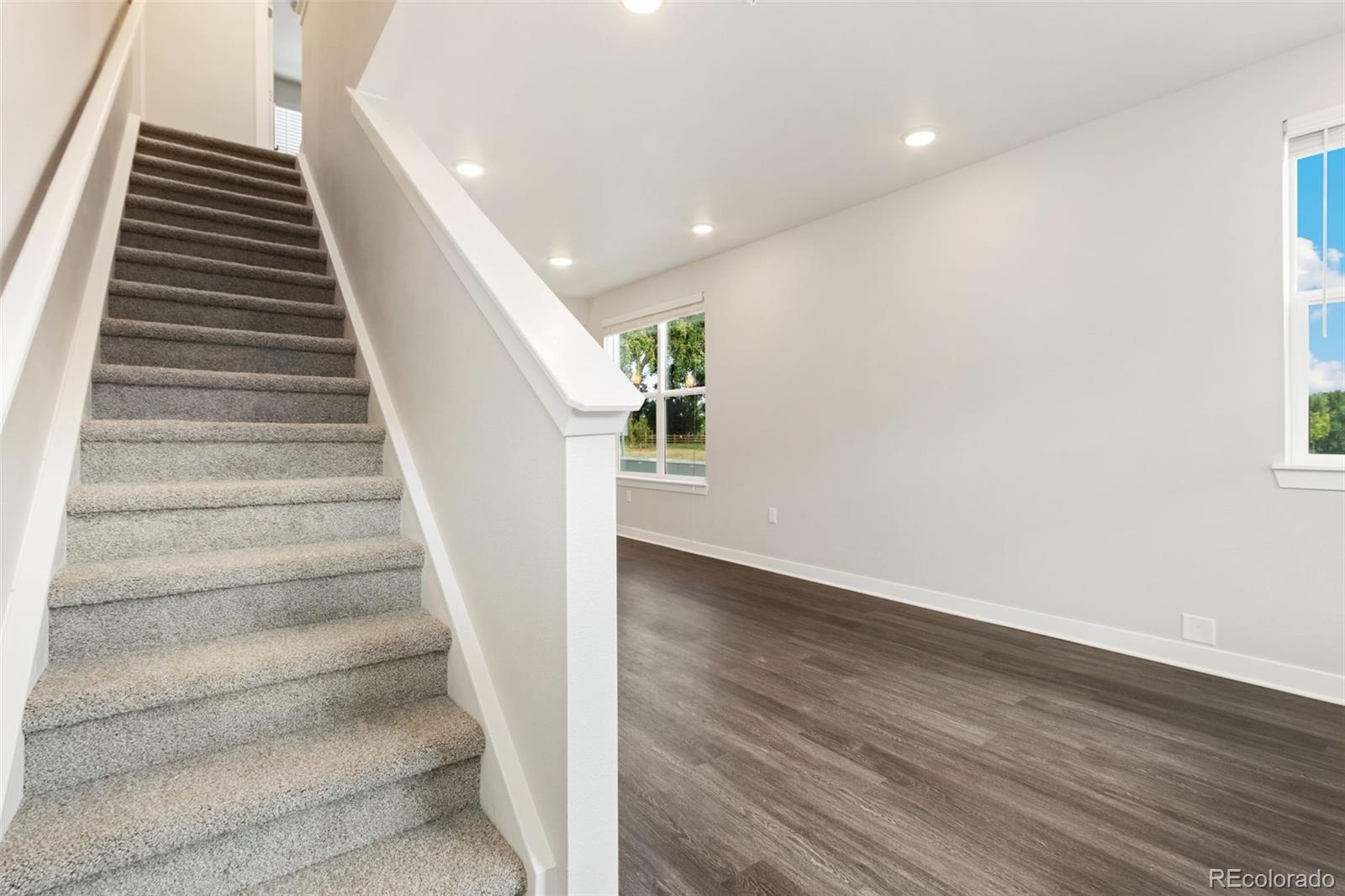 Carpeted staircase beside a bright, vacant room with dark wood-look flooring.