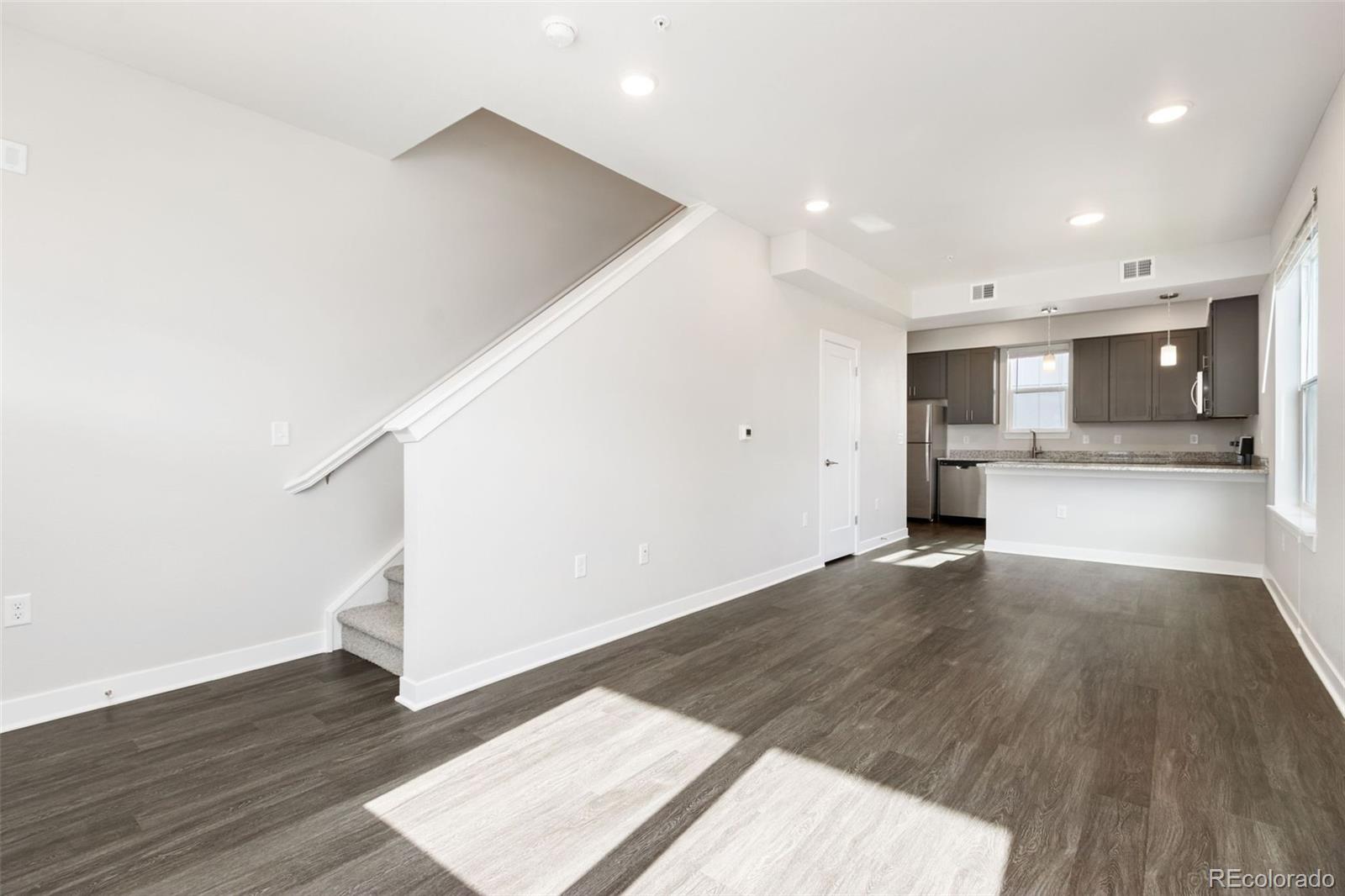 Vacant living room opens to a kitchen, with a carpeted staircase and sunlight across dark flooring.