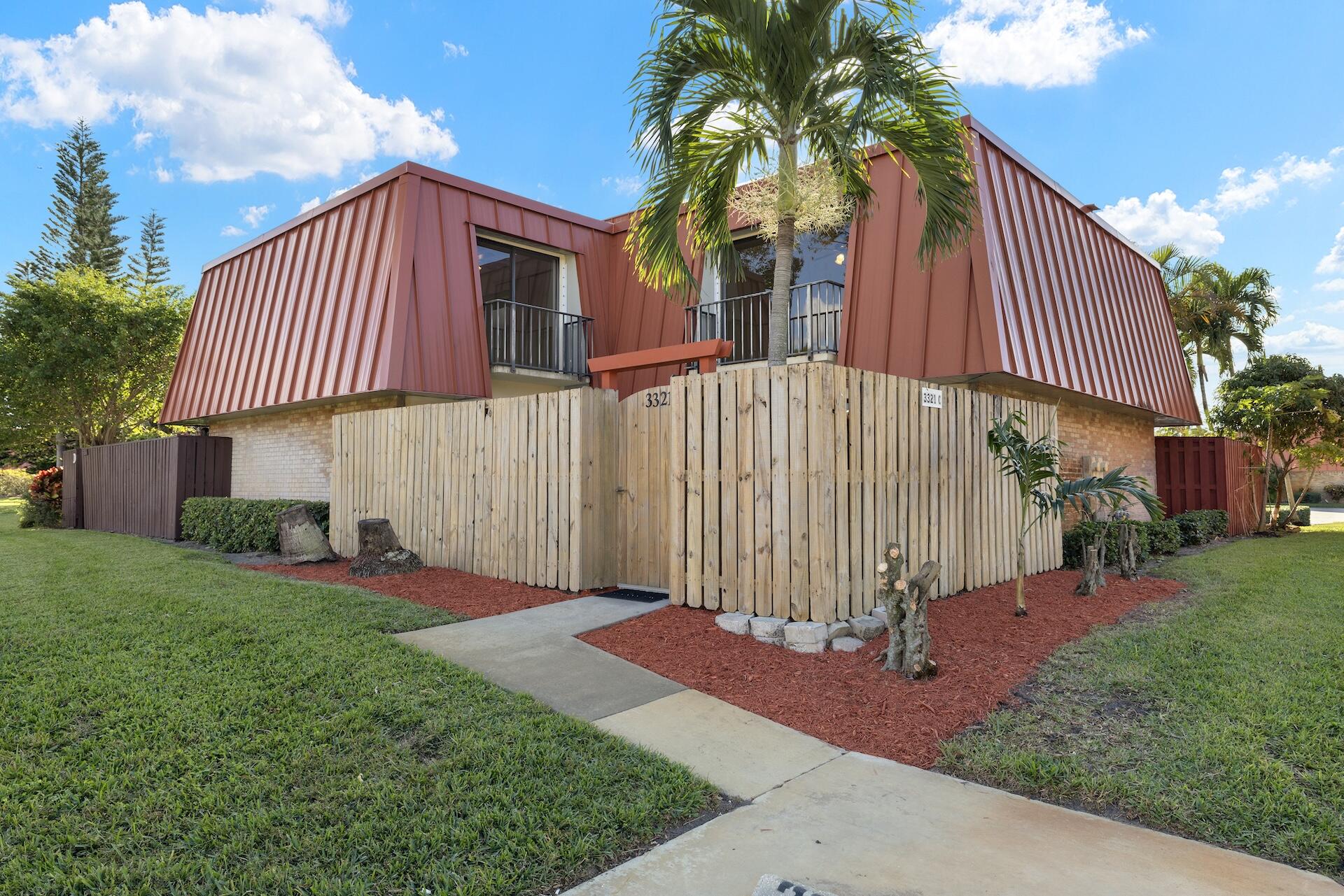 Two-story home with red metal upper siding, balconies, a wooden fence, and a palm tree.