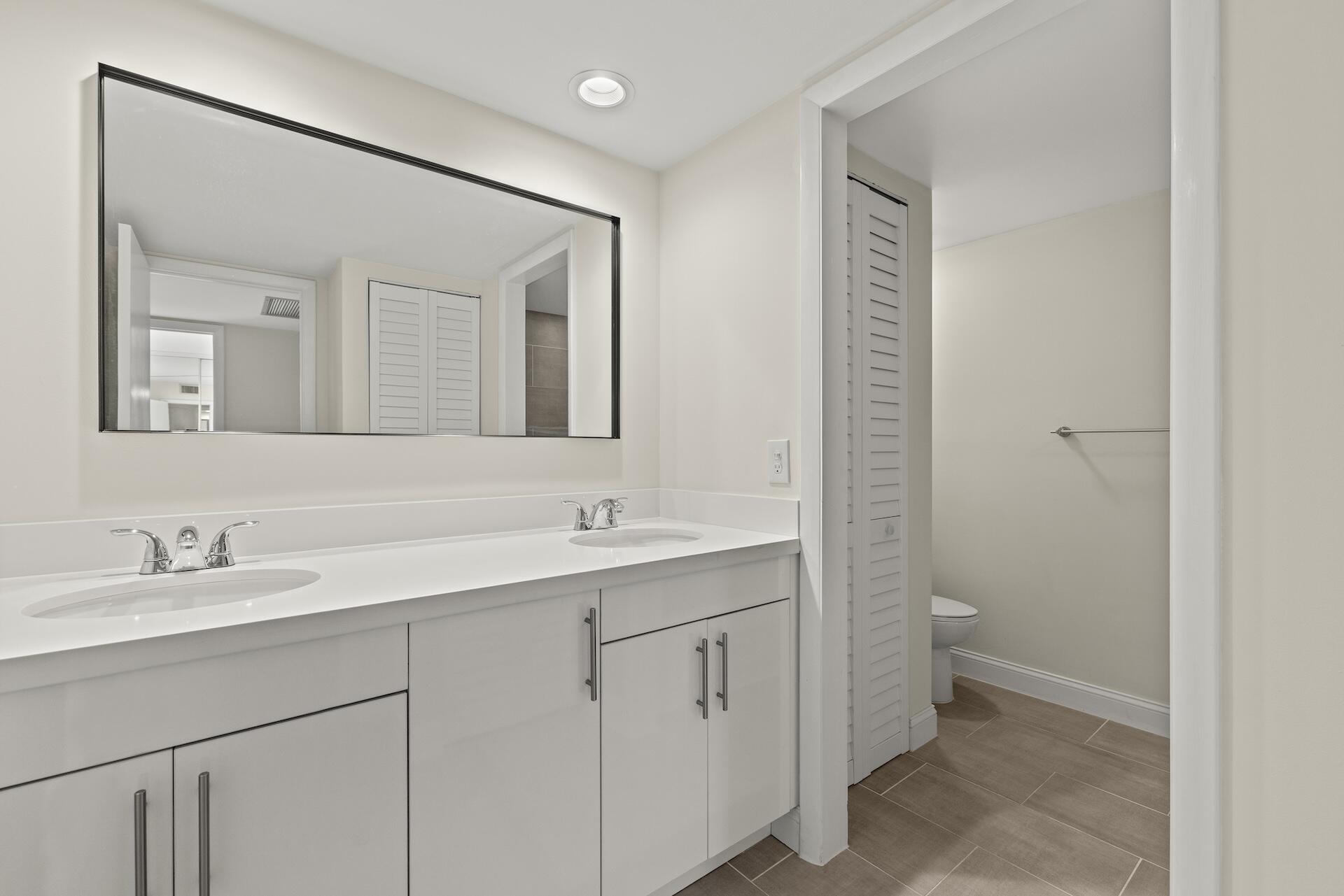 Bathroom with a long double-sink vanity and mirror beside an adjoining toilet and shower area.