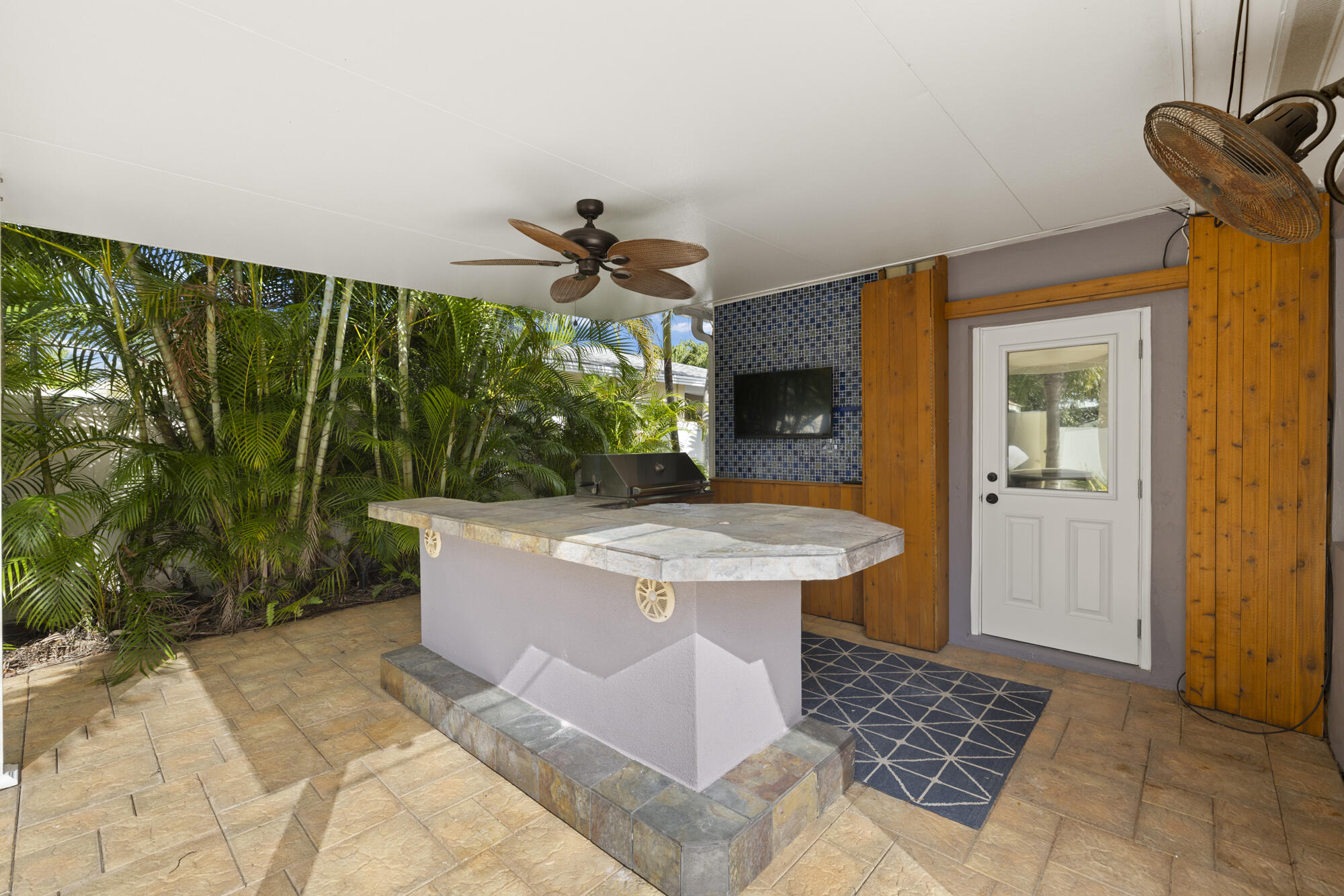 Covered outdoor kitchen with stone island, built-in grill, mosaic-tile TV wall, and tropical palms.