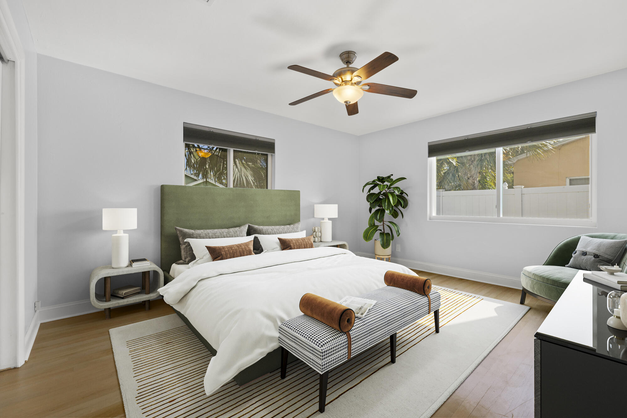Furnished bedroom with an olive-green headboard, patterned bench, and windows overlooking a fenced yard.
