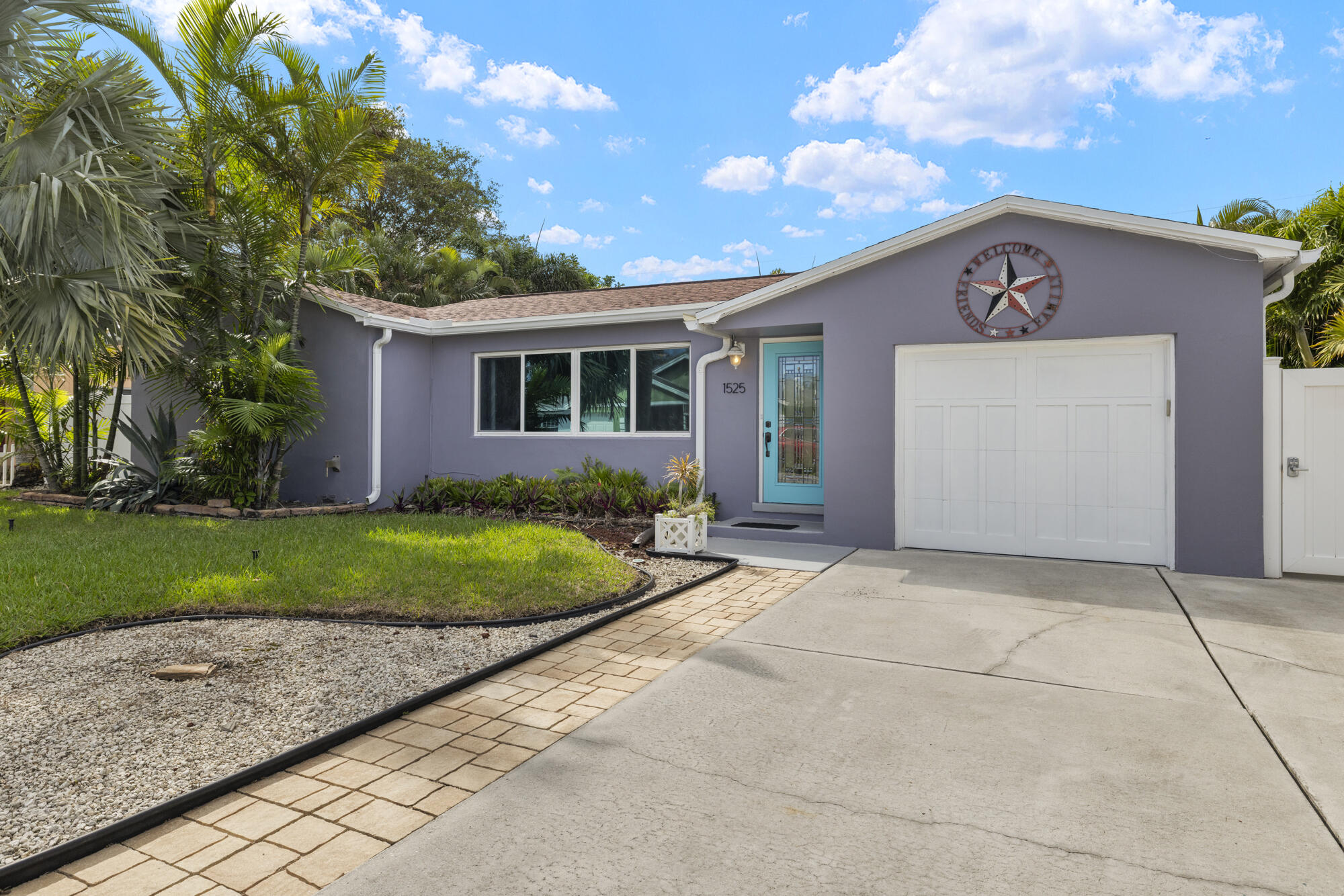Single-story purple-gray house with a turquoise front door, white garage, and tropical landscaping.