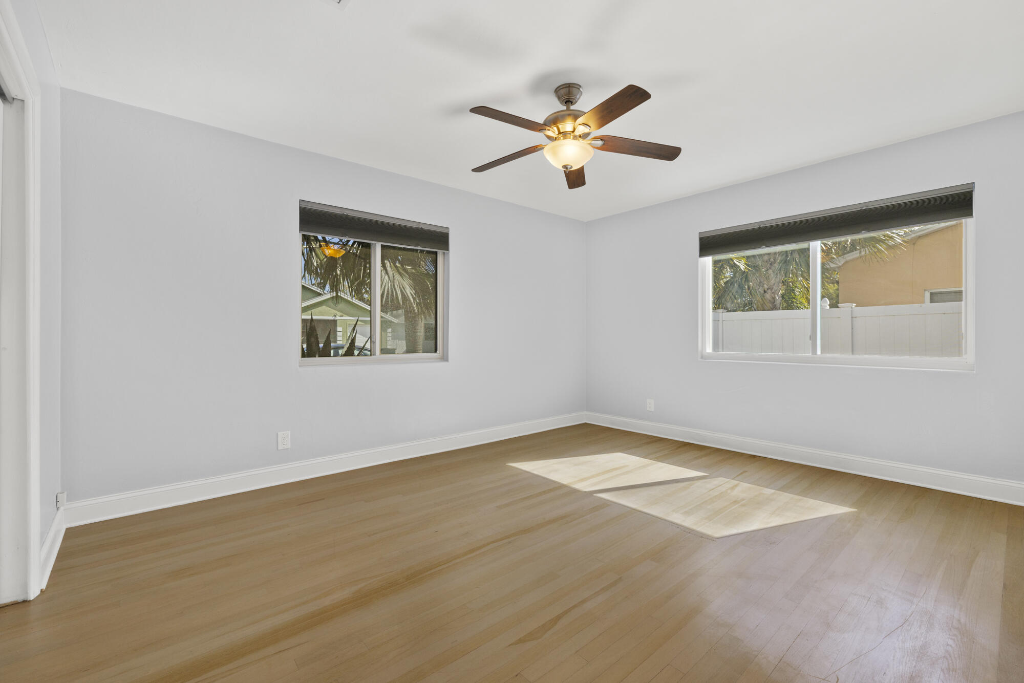 Empty bedroom with light wood floors, two windows, and a ceiling fan with a lit fixture.