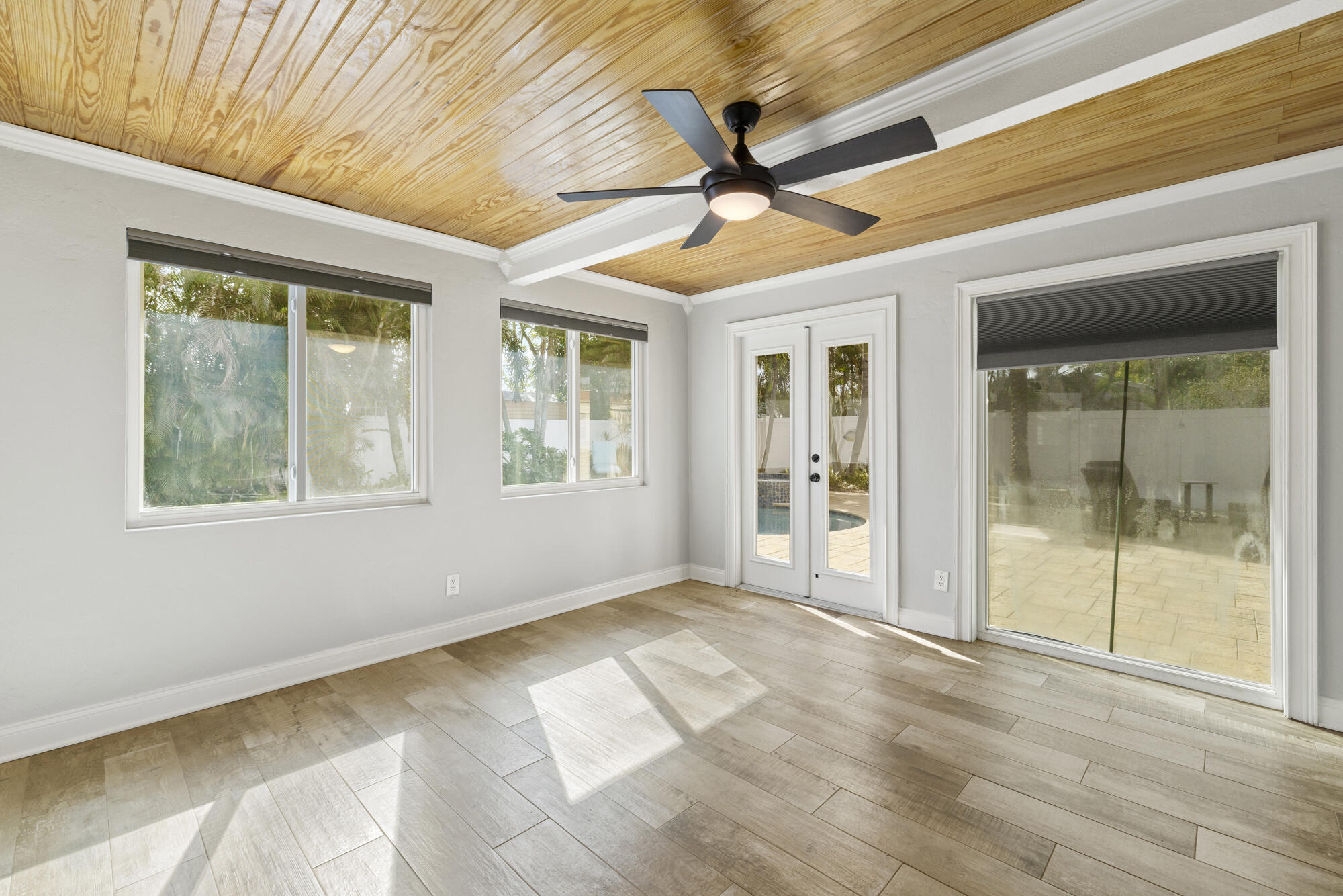 Empty sunroom with a wood-plank ceiling, ceiling fan, and glass doors overlooking a patio and pool.