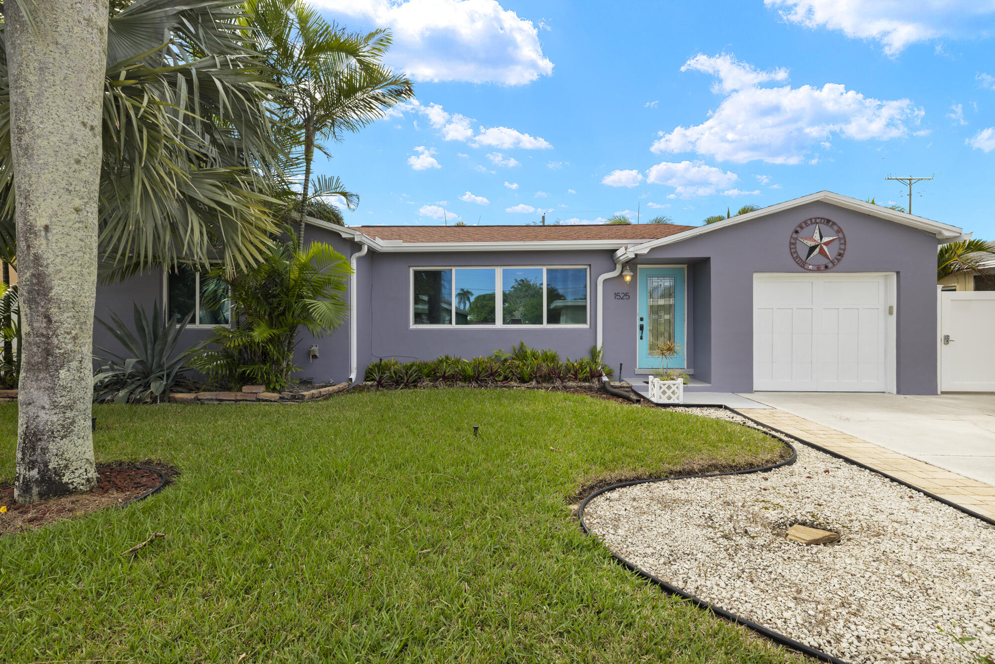 Single-story purple-gray home with a turquoise front door, white garage, lawn, and palm landscaping.