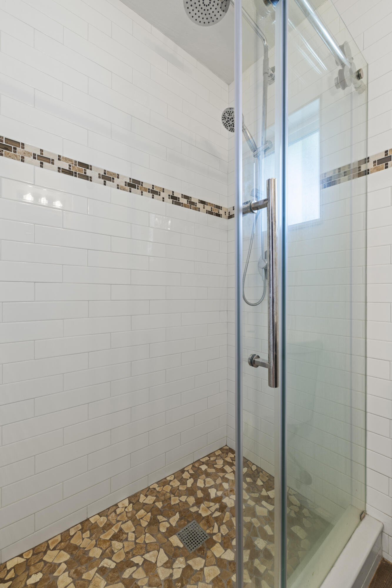 Walk-in shower with glass doors, white subway tile, mosaic accents, and a stone-patterned floor.
