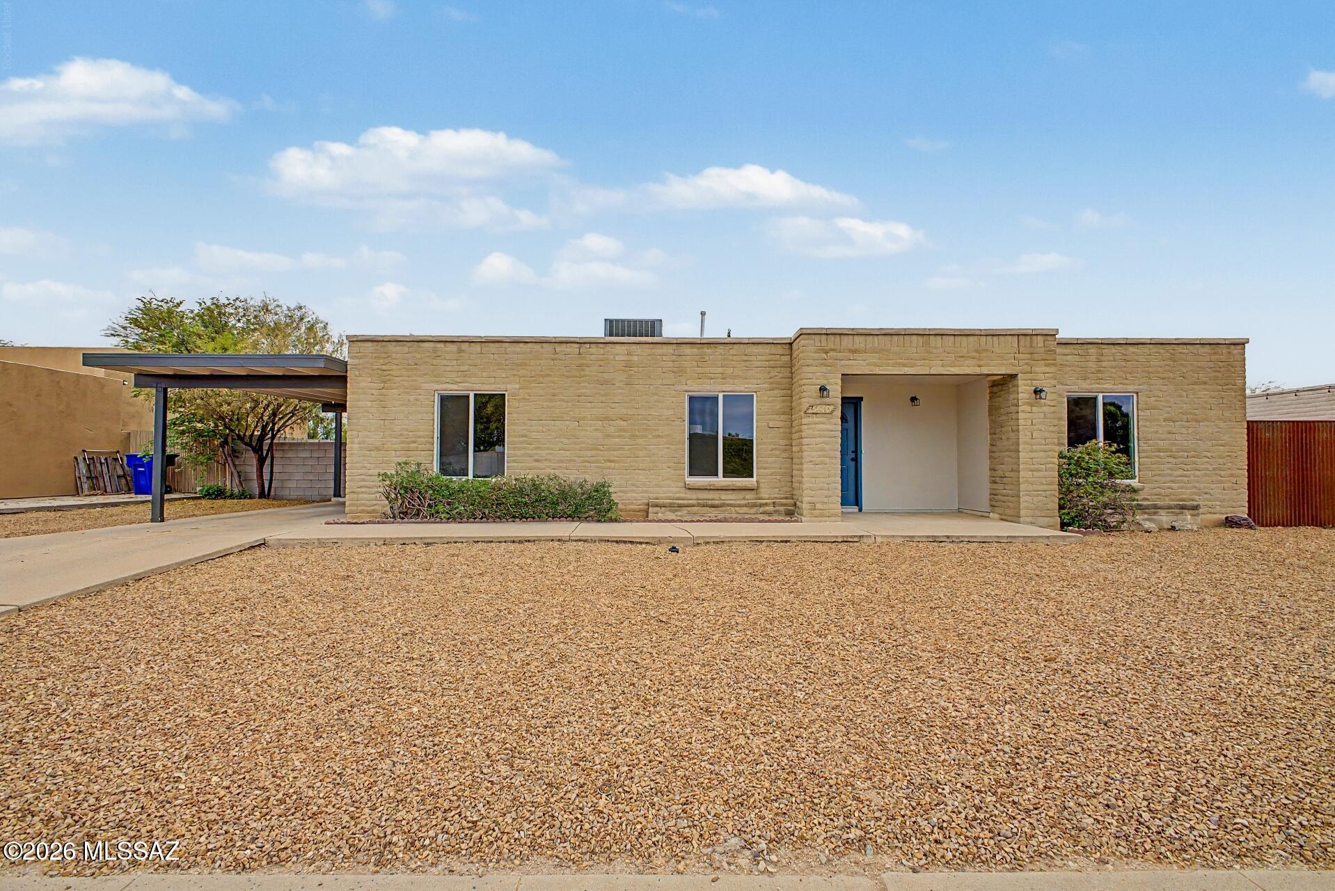 Single-story tan masonry home with a recessed blue entrance, covered carport, and gravel front yard.
