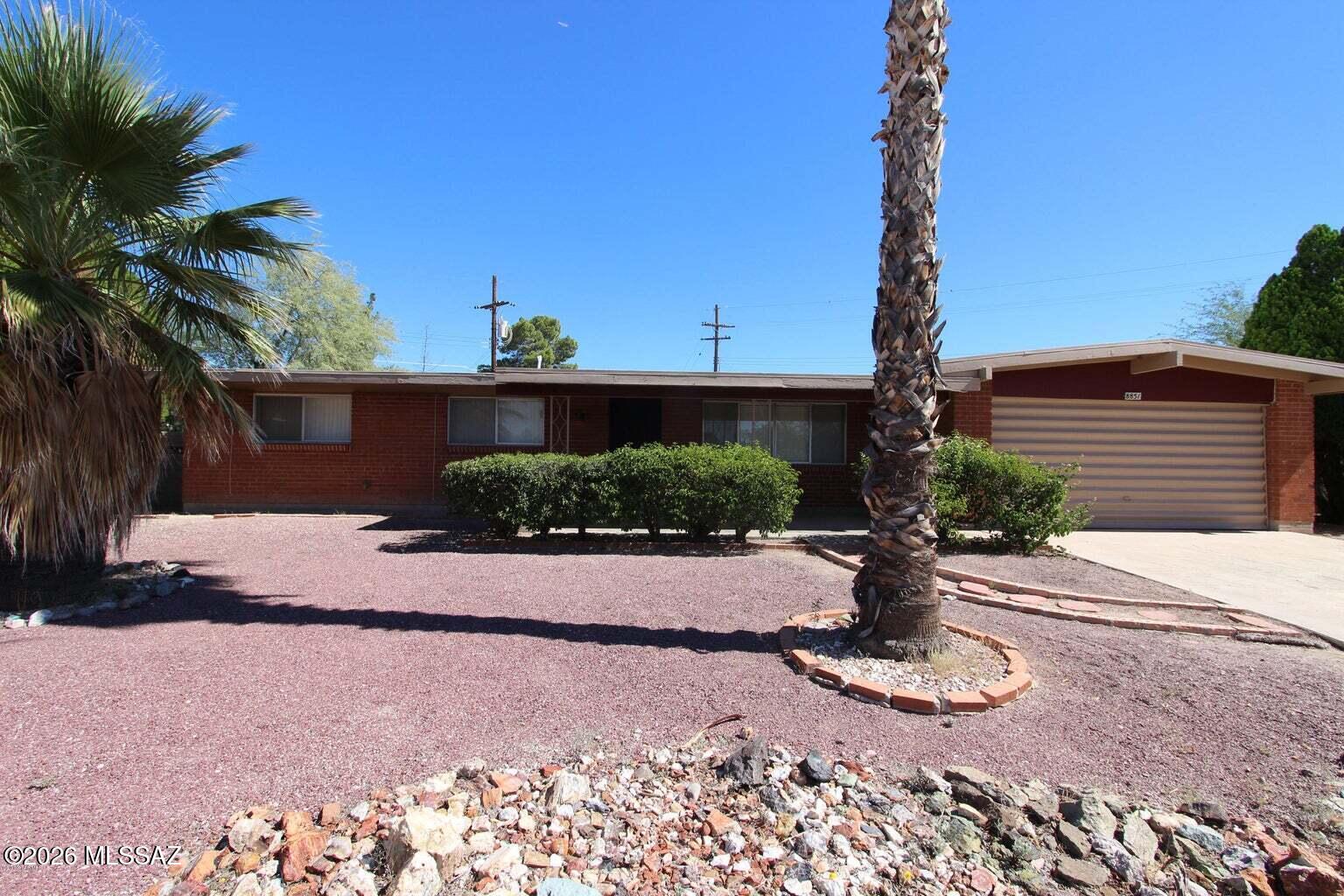 Single-story brick ranch house with a garage, gravel yard, trimmed shrubs, and palm trees.