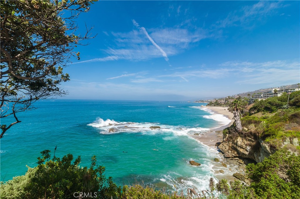 Coastal ocean view with waves, a sandy beach, rocky cliffs, and homes along the hillside.