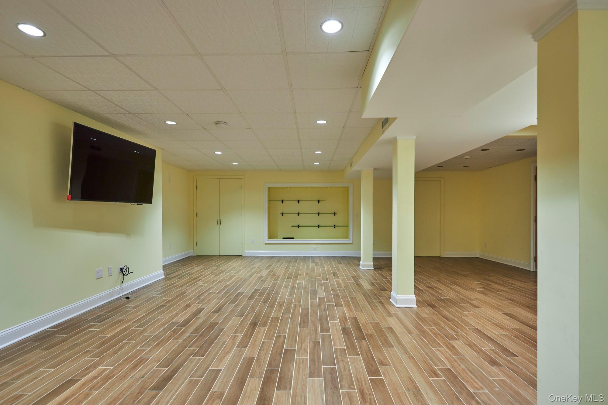Vacant recreation room with wood-look flooring, wall-mounted television, and recessed display shelves.