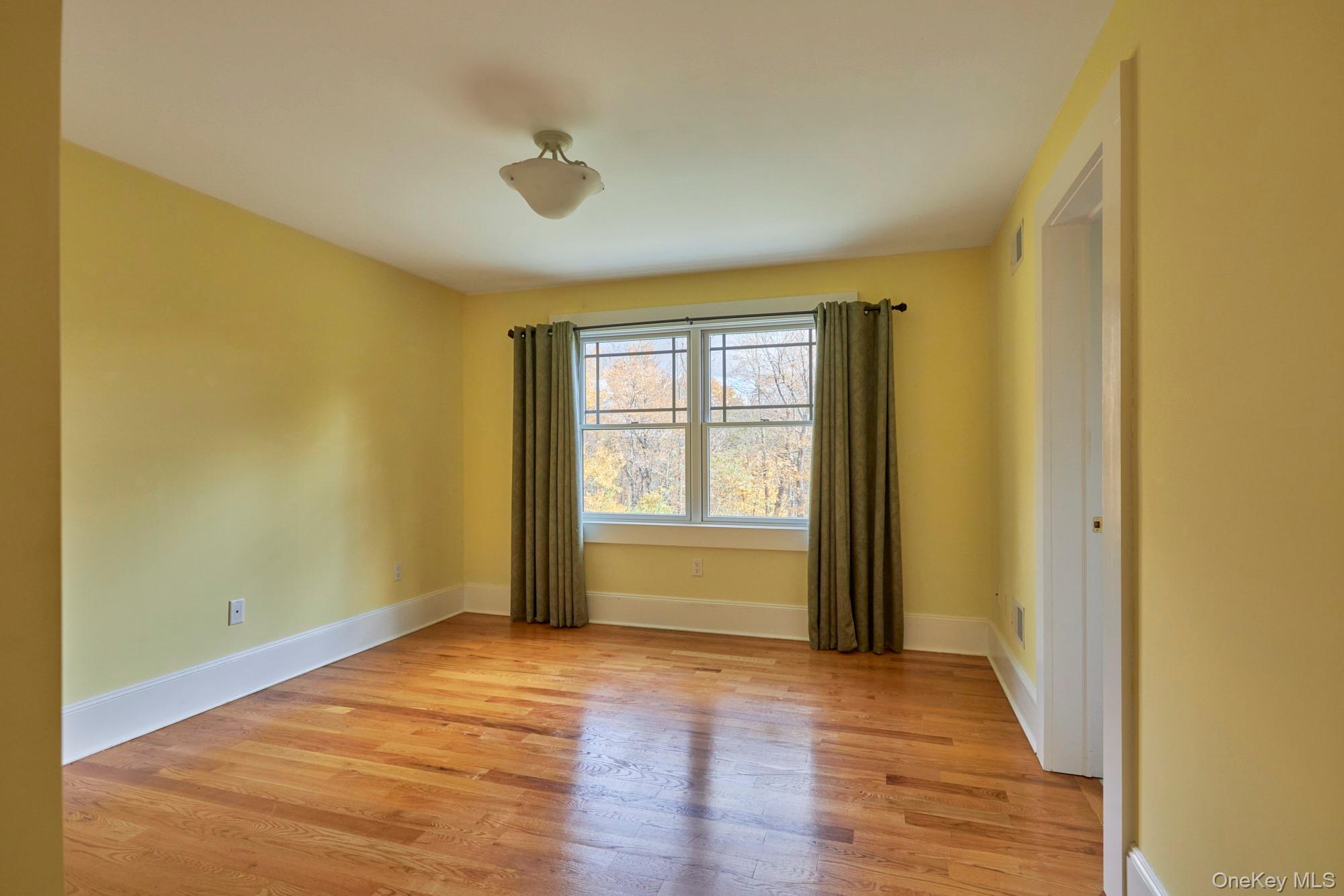 Empty room with yellow walls, hardwood floors, and a curtained window overlooking autumn trees.