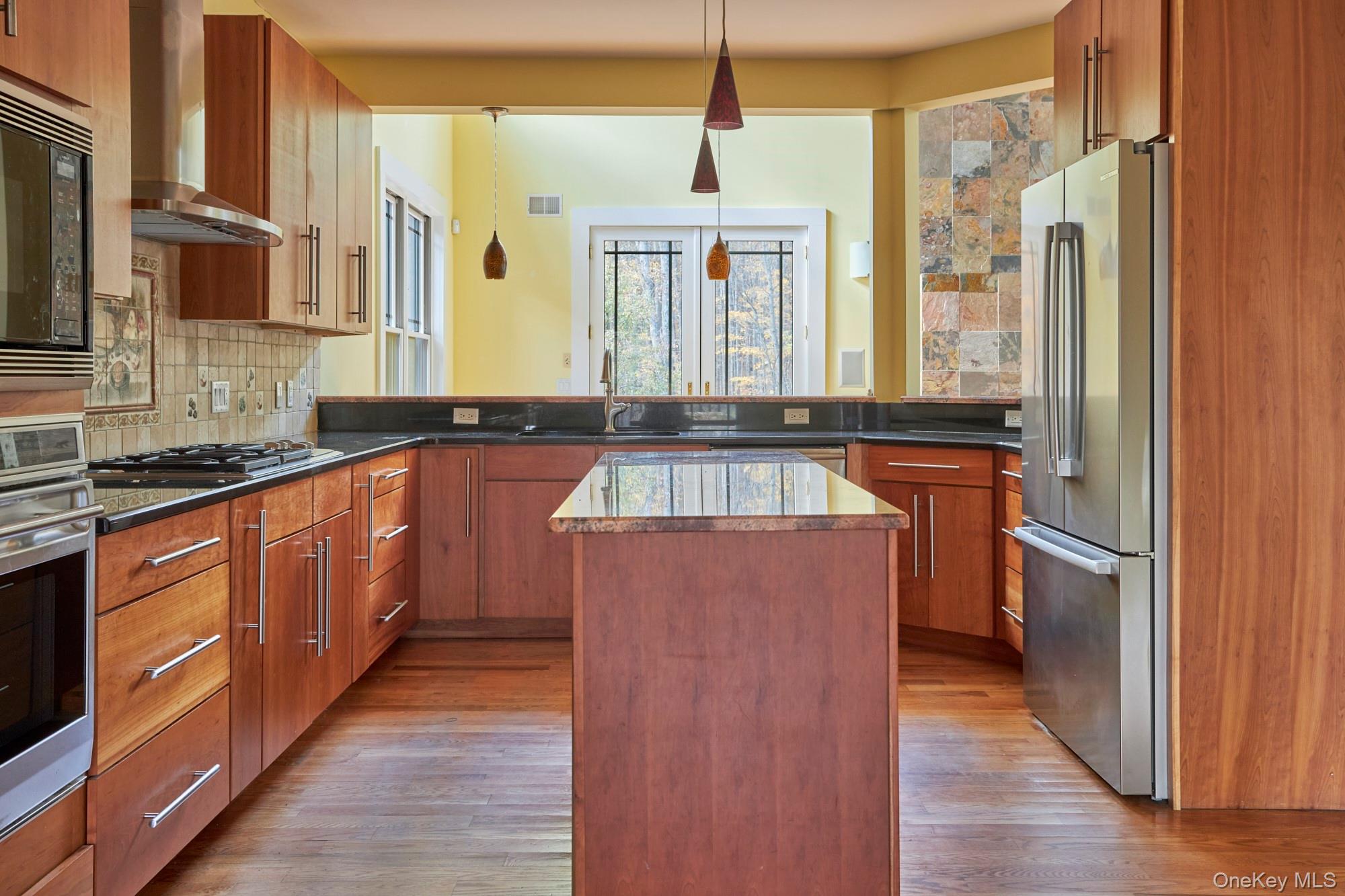 Modern kitchen with warm wood cabinetry, a central island, stainless steel appliances, and windows overlooking autumn trees.