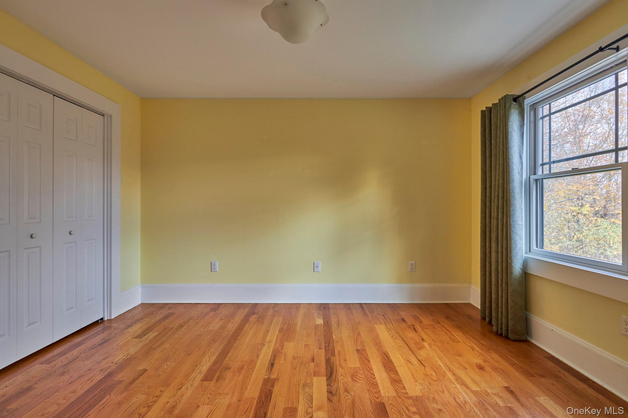 Empty yellow room with hardwood floors, closet doors, and a large window overlooking autumn trees.