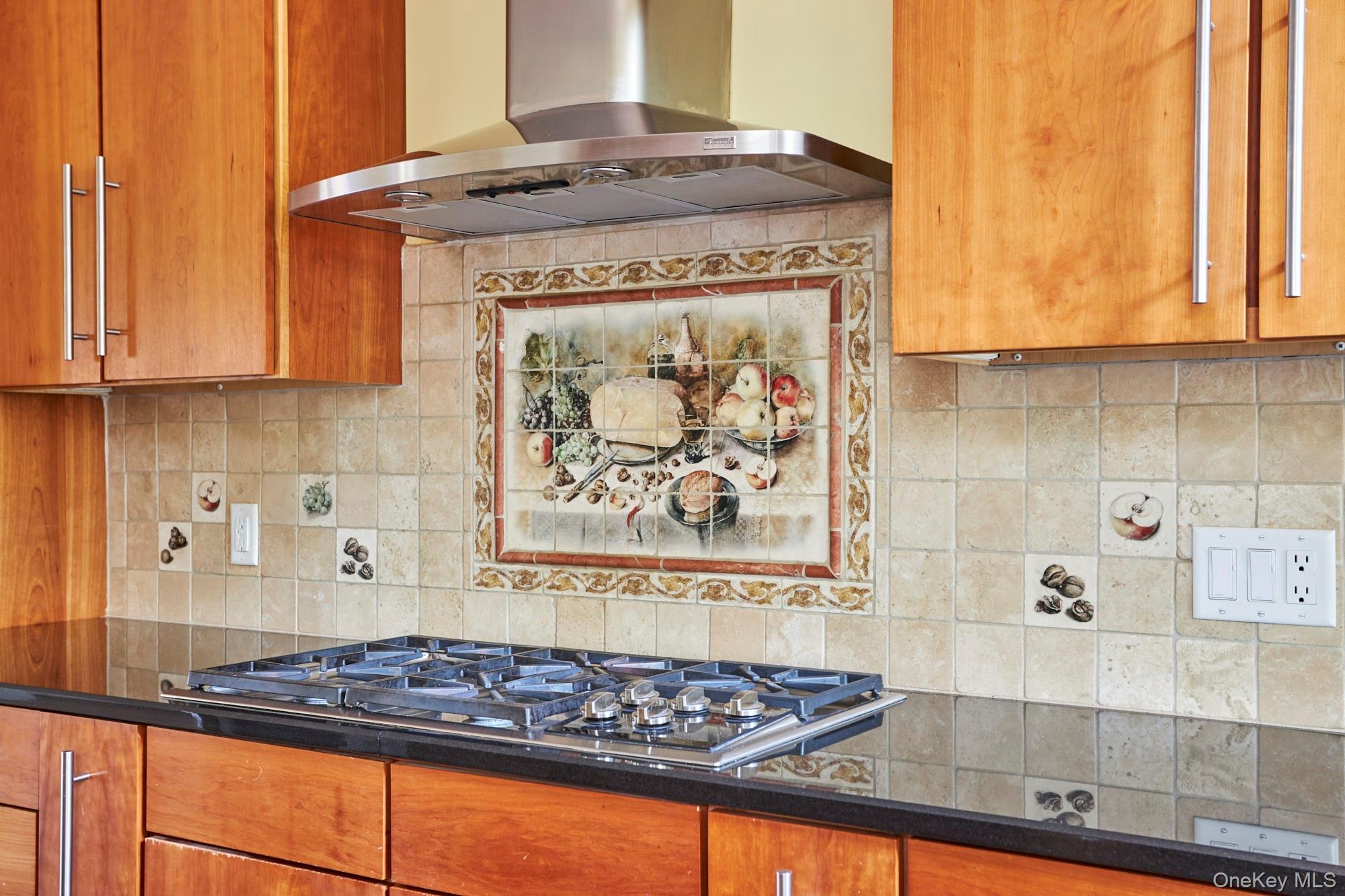 Kitchen with wood cabinets, a stainless steel gas cooktop, and a decorative fruit-pattern tile backsplash.