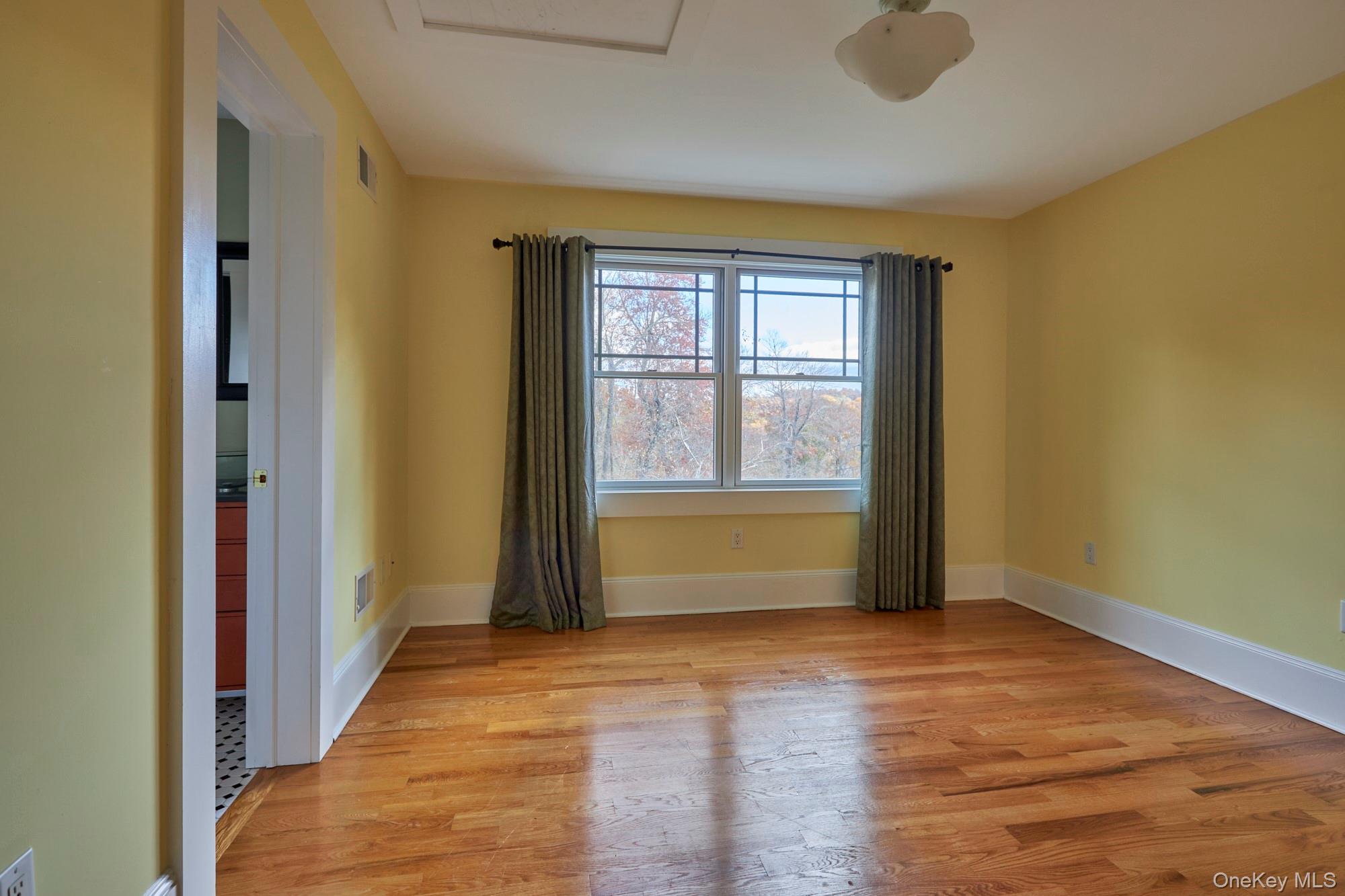 Empty yellow-walled room with hardwood floors and a curtained window overlooking autumn trees.