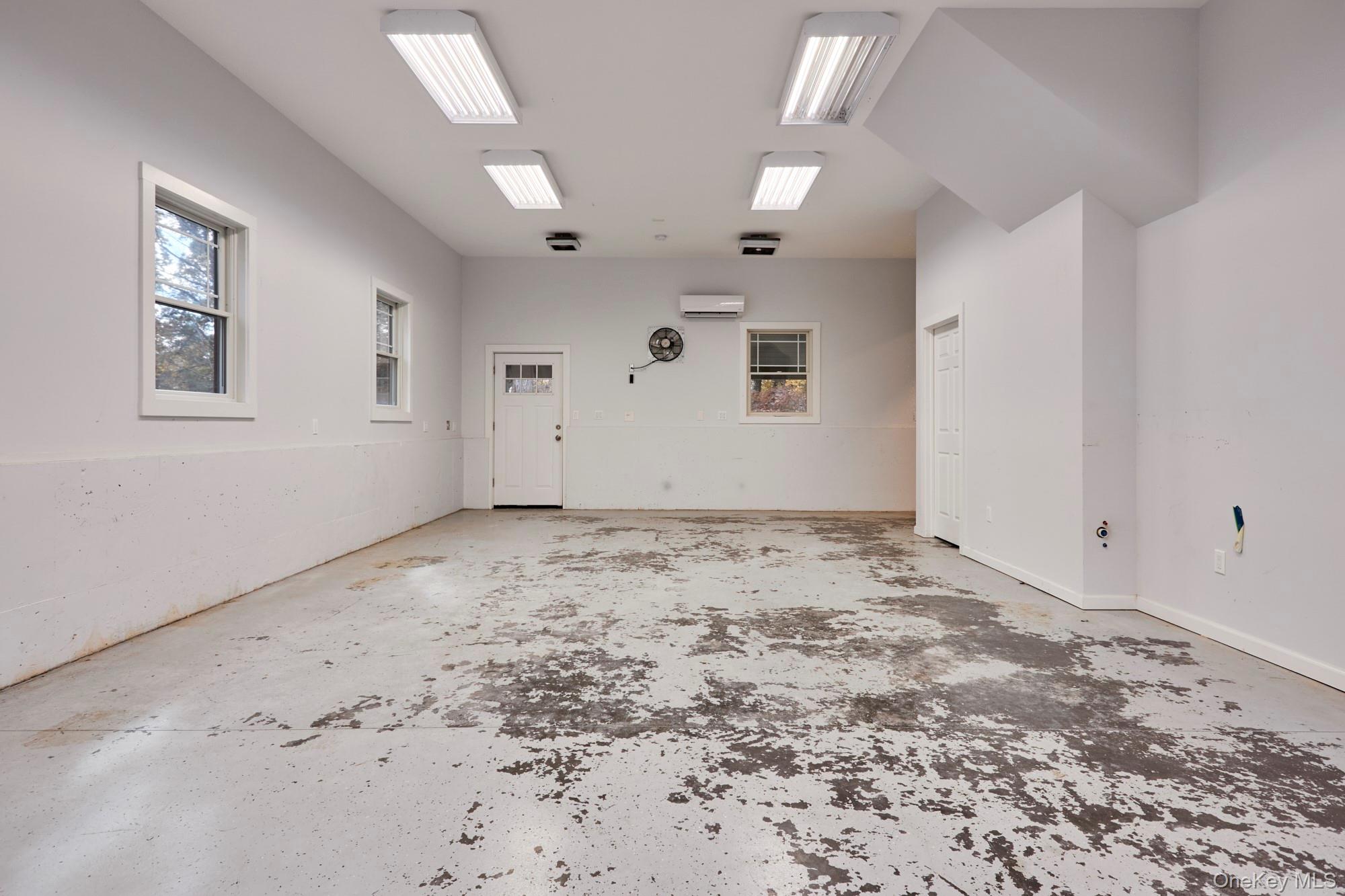 Empty garage with a heavily worn concrete floor, fluorescent ceiling lights, and several windows.