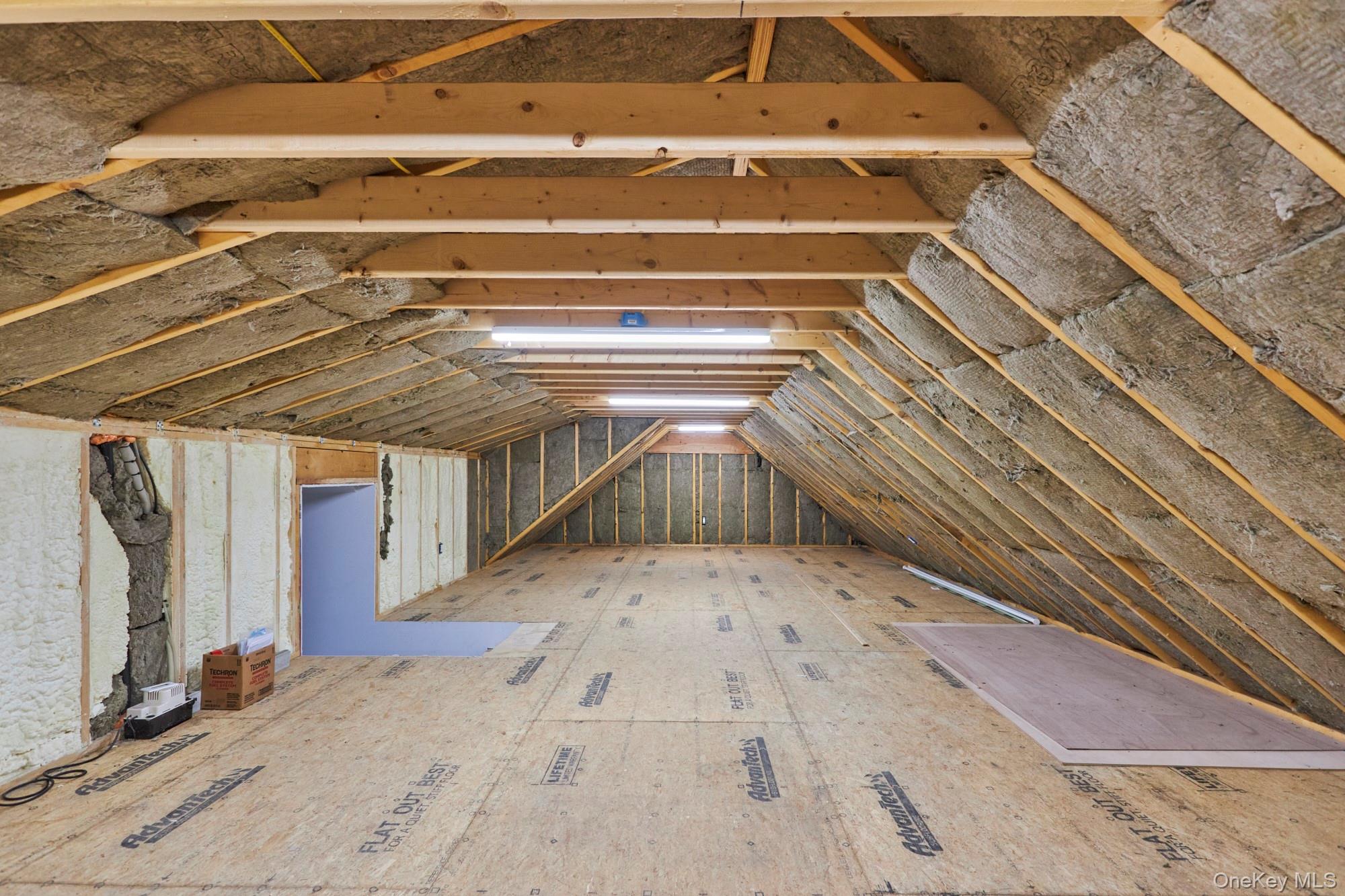 Unfinished attic with exposed rafters, sloped roof insulation, plywood flooring, and fluorescent lights.