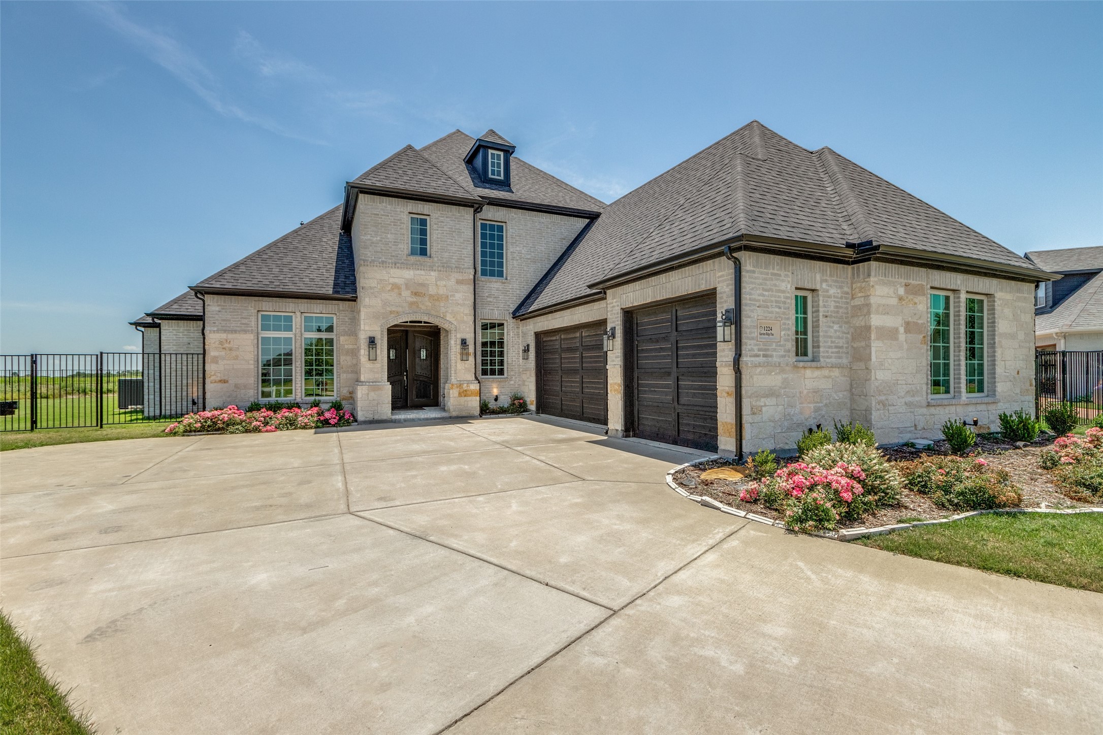Two-story stone house with dark garage doors, a wide driveway, and pink flower beds.